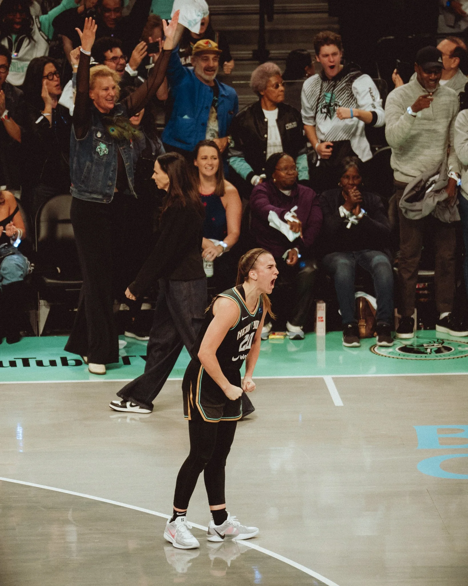 A person in a black basketball uniform standing on a basketball court, celebrating with clenched fists. Behind her, a basketball game is happening with spectators and players showing excitement.