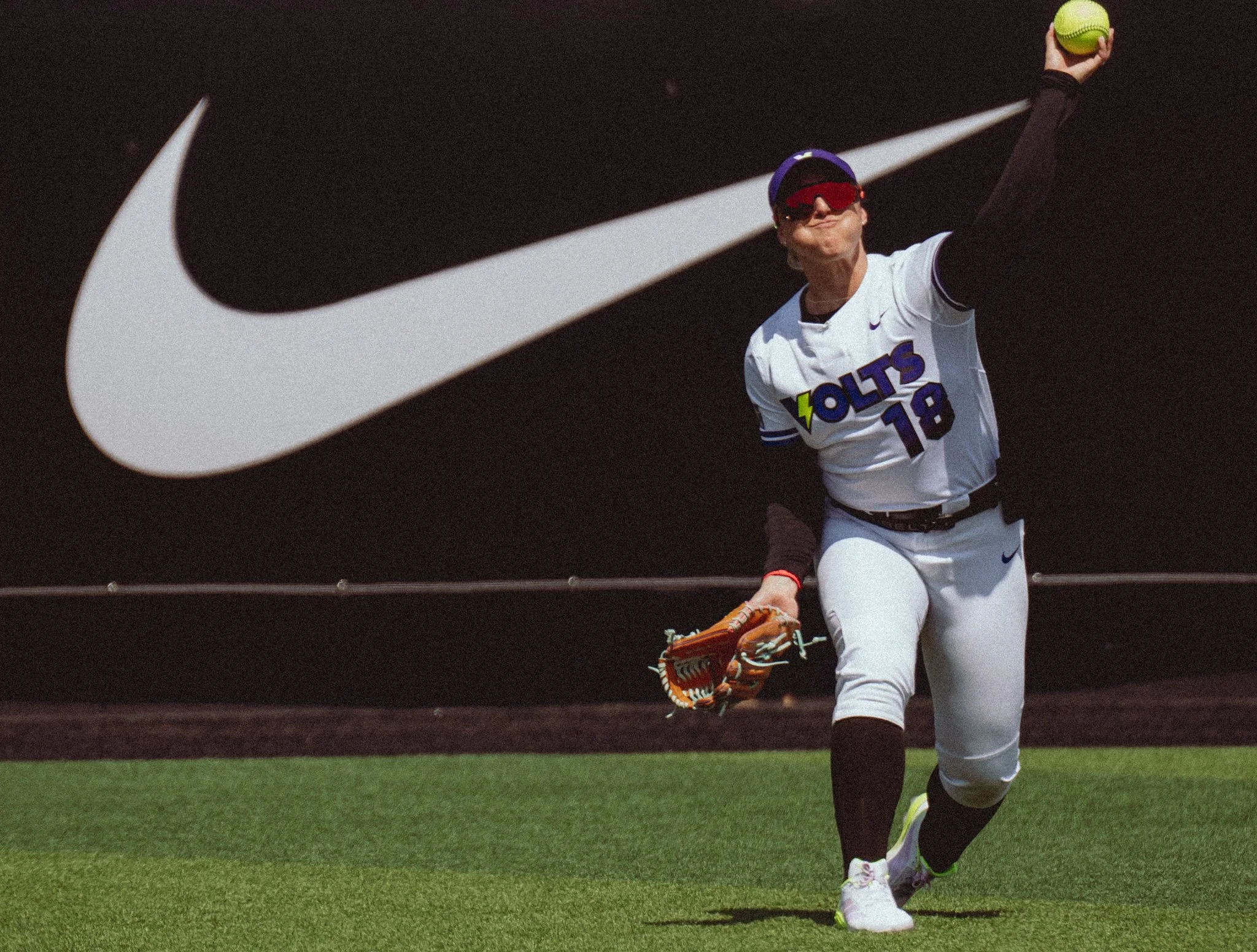 A woman baseball player in a white uniform with purple and yellow accents catching a ball with a baseball glove on her left hand. She is wearing sunglasses, a cap, and has a serious expression. She is on a field with a green grass infield and stands 