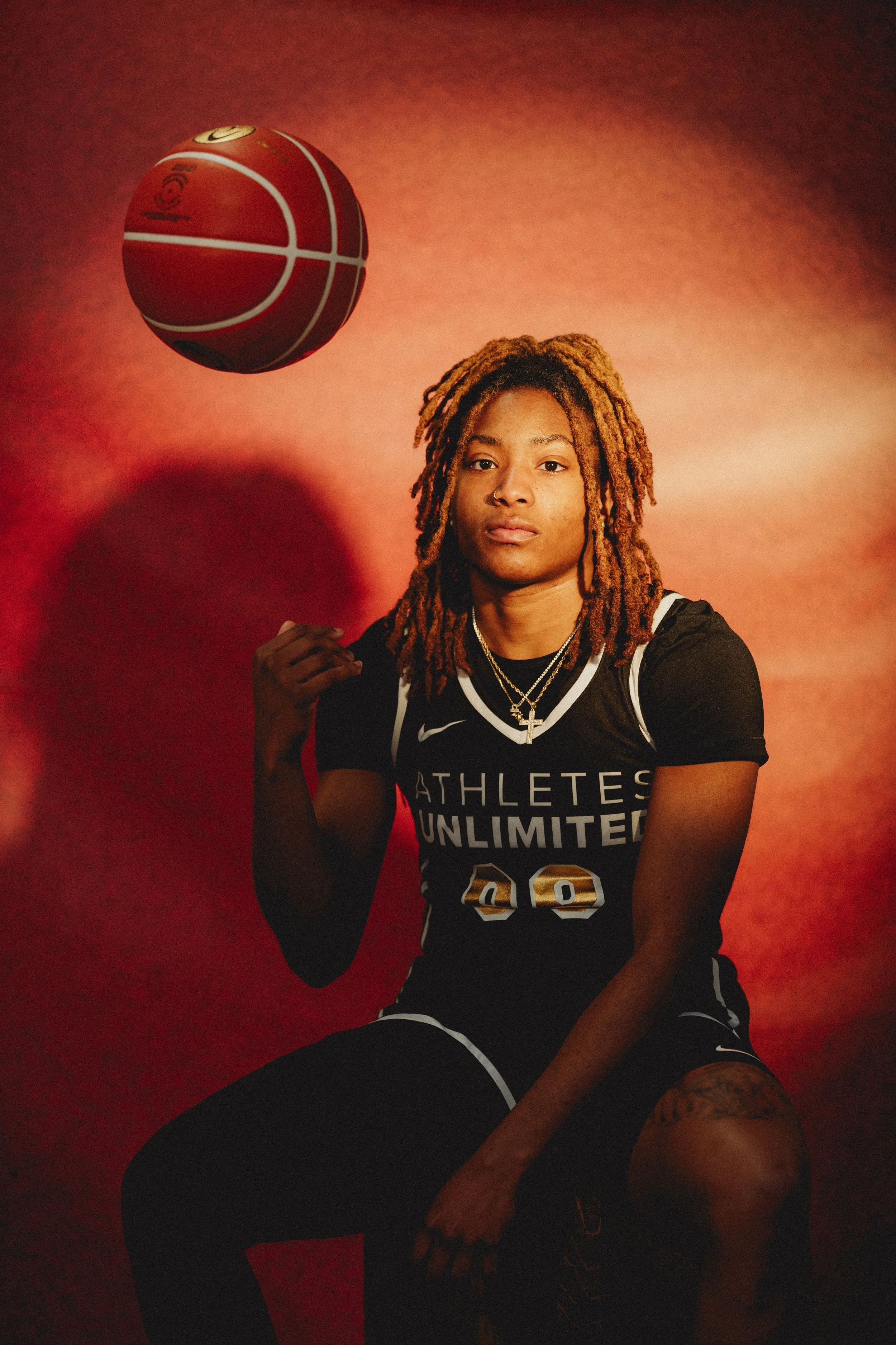 A woman with long blonde dreadlocks wearing a black sports jersey with the words 'ATHLETES UNLIMITED' and the number 28, sitting with one knee up, against a reddish-brown background. She is looking directly at the camera with a focused expression, an