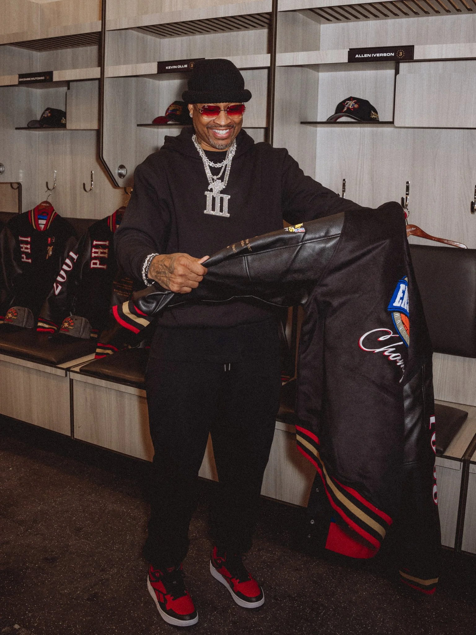 A man in a black hoodie, black pants, red and black sneakers, and red sunglasses is smiling and holding a black jacket in a sports locker room with caps and jackets on display.