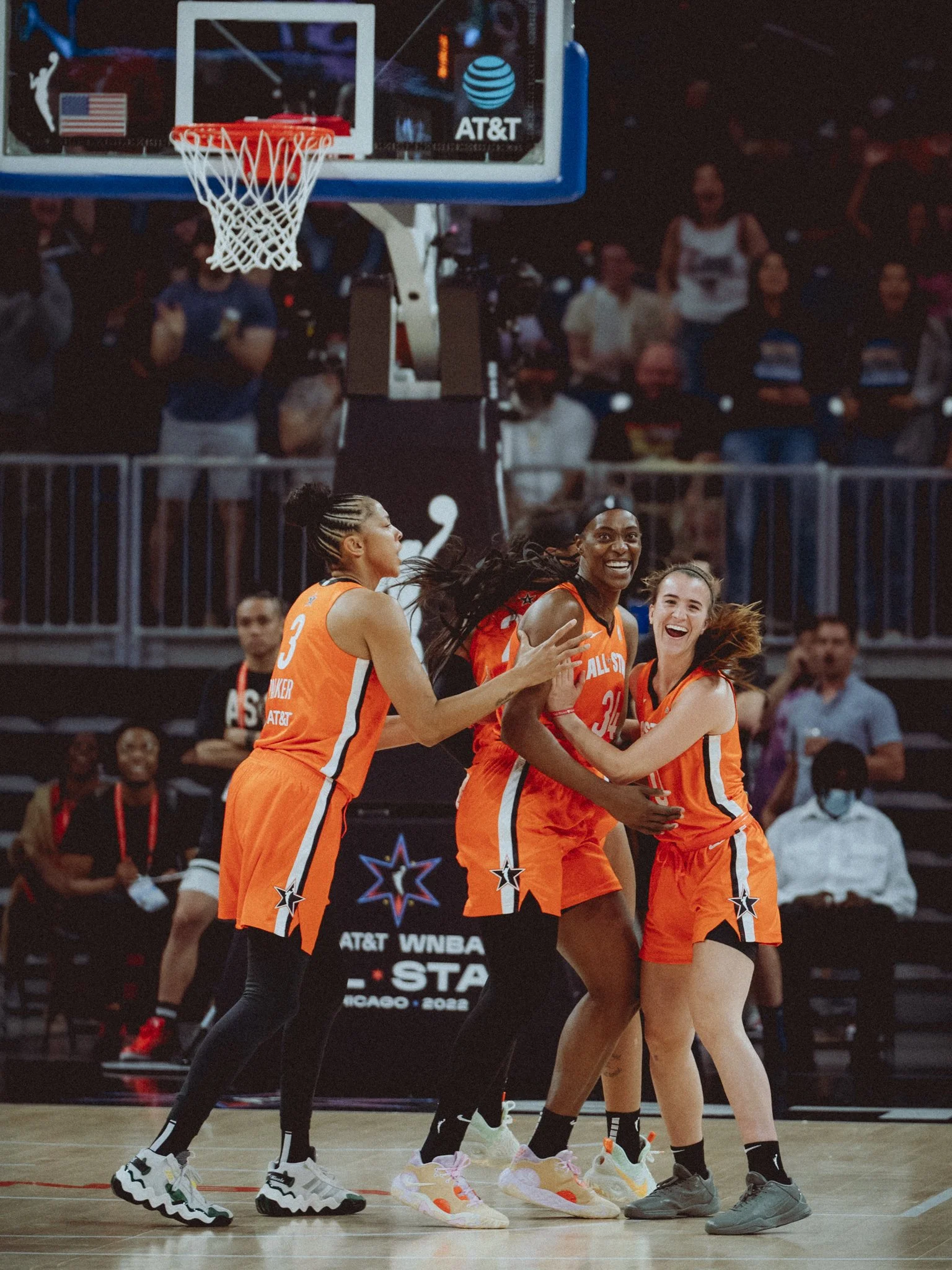 Women basketball players wearing orange jerseys celebrating on a basketball court with a basketball hoop overhead and spectators in the background.