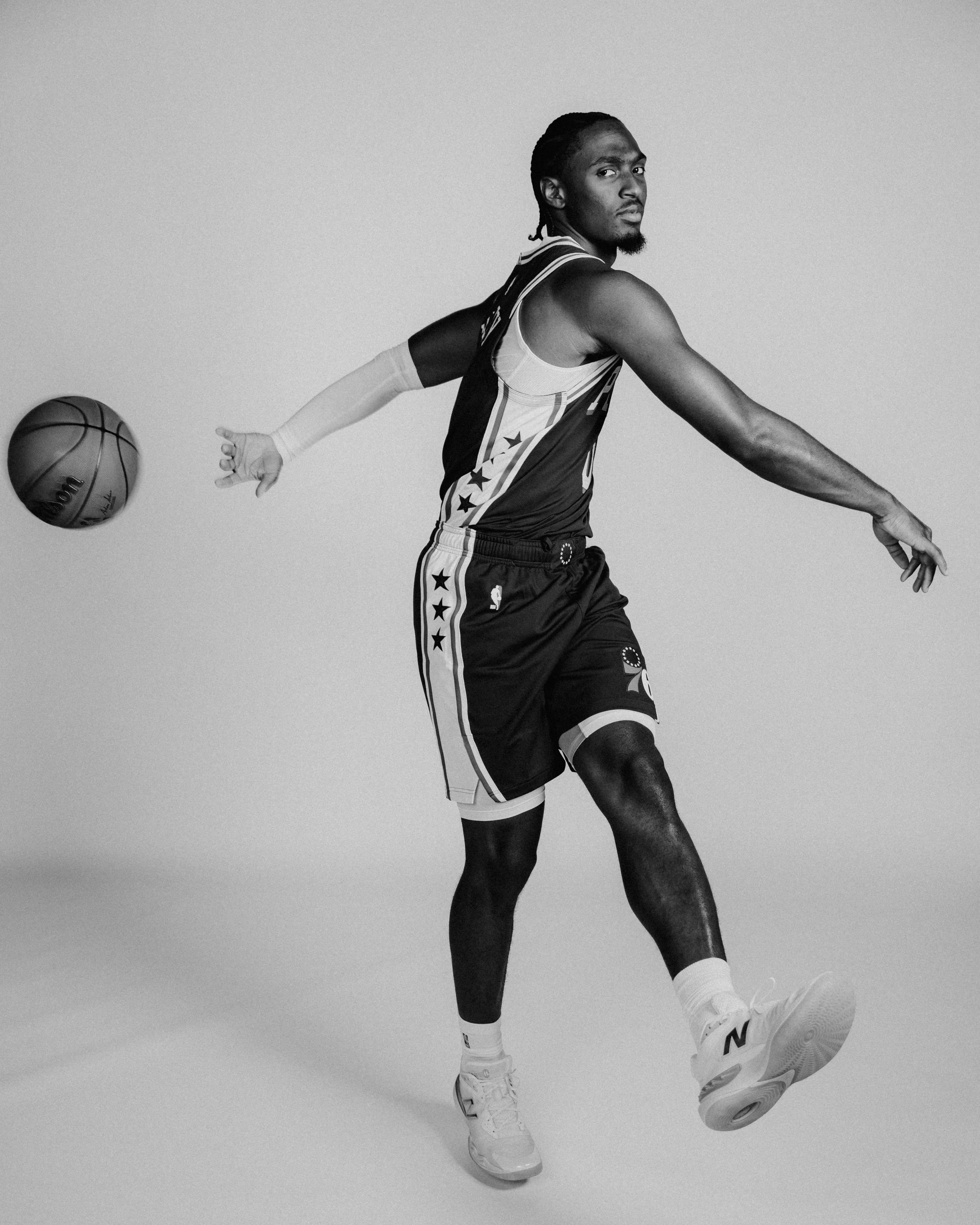 Black and white photo of NBA player, VJ Edgecombe in a sports jersey and shorts, performing a move on the court with a basketball in one hand. He is looking back over his shoulder, with one leg raised and the other extended.