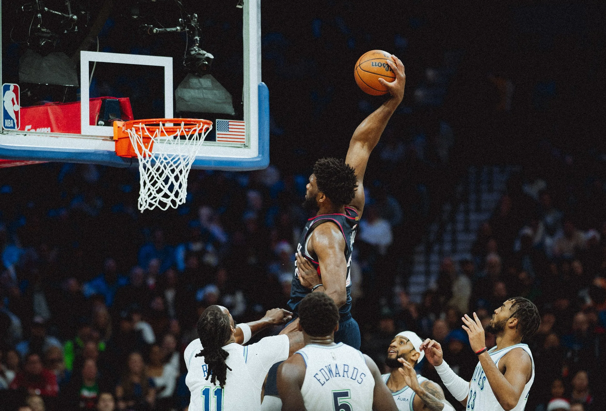 A basketball player in a black uniform is jumping to make a shot at the hoop. Several players in white uniforms are reaching up towards him, and the crowd is visible in the background.