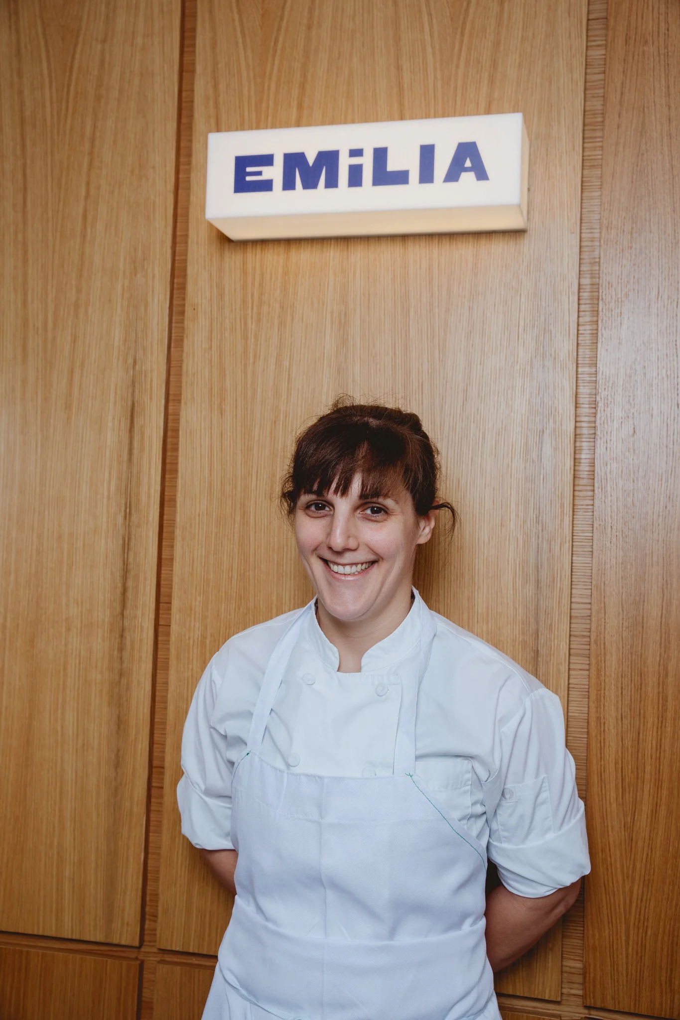 A woman dressed as a chef standing in front of wooden cabinets with a sign that reads 'EMILIA' above her head.