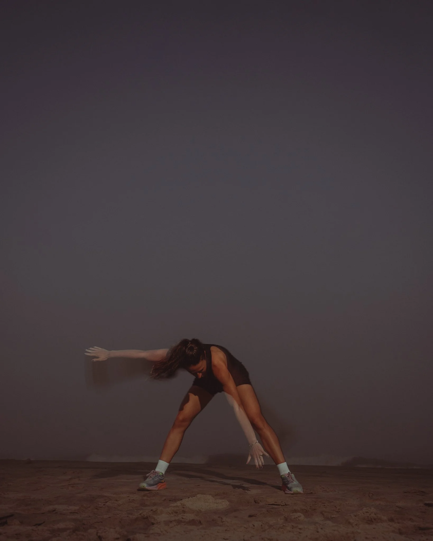 A woman in athletic clothing is doing a physical exercise, bending forward with one arm extended to the side and the other arm reaching toward the ground, on a sandy surface at night with a dark sky in the background.