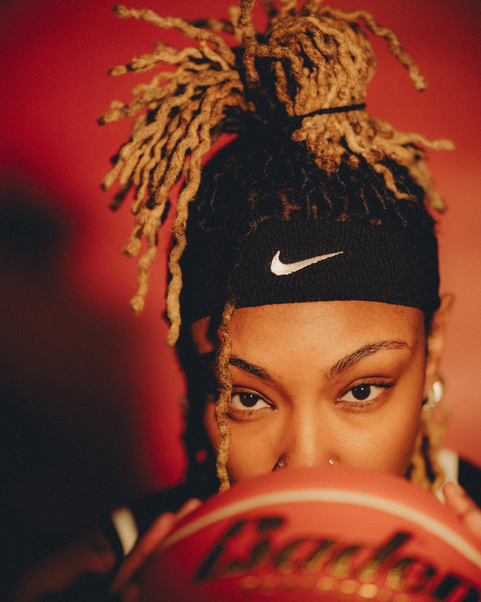 A woman with dreadlocks and brown skin peeks over a spiral notebook. She is wearing a black sock cap with a white Nike logo and has a septum piercing. The background is red and slightly blurred, focusing on her face.