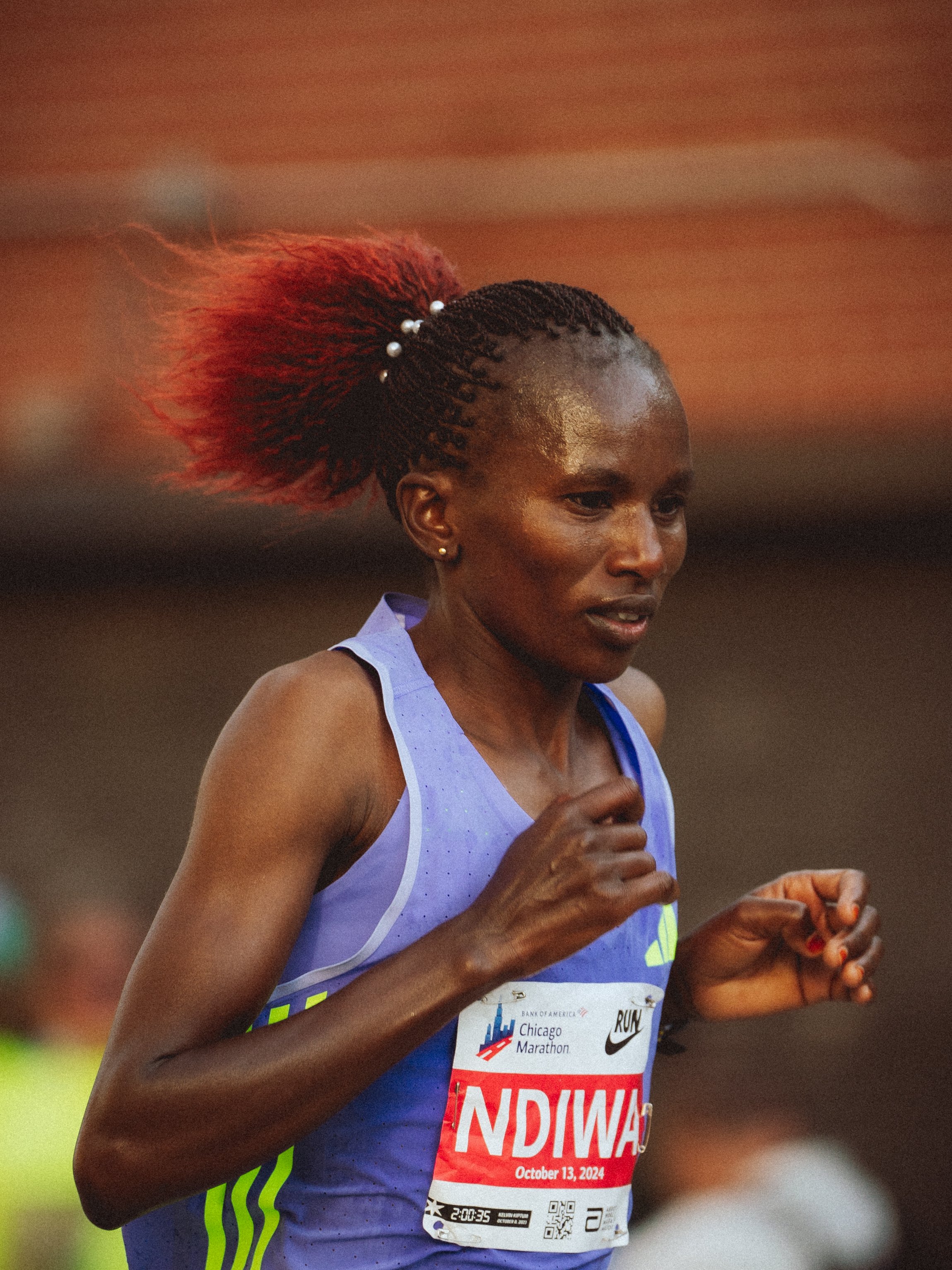 A woman running in a marathon, wearing a blue athletic tank top with green accents, a race bib with 'INDIANA' and the date October 13, 2024, and a watch on her left wrist. She has braided hair with red extensions and white beads.