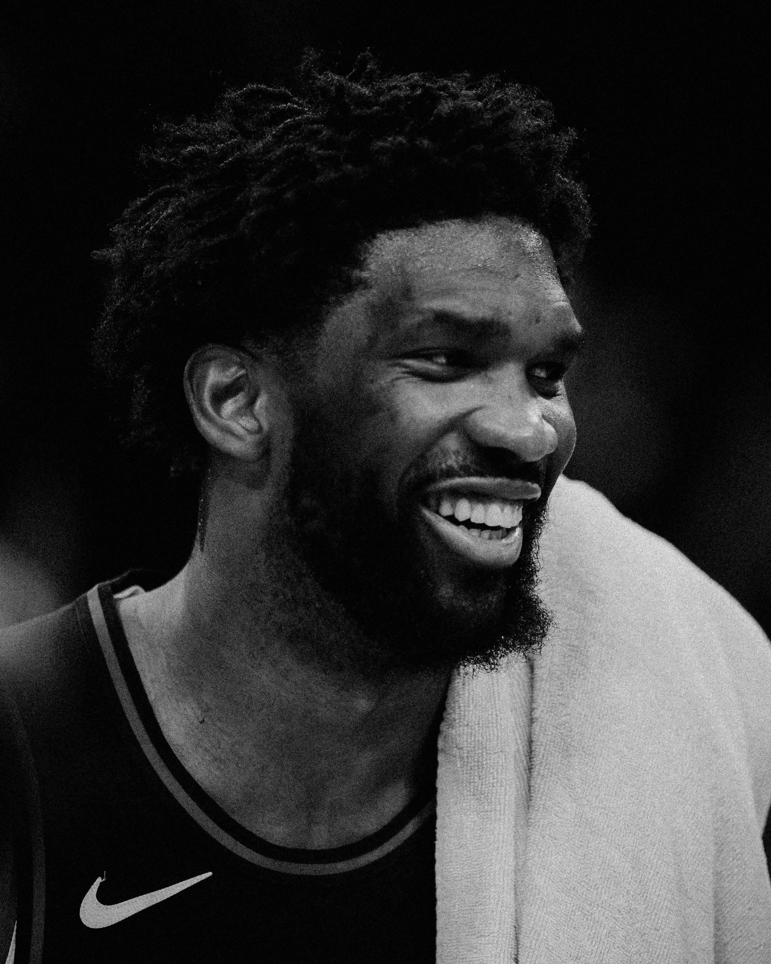 Close-up black and white photo of a man smiling with a beard, wearing a Nike shirt, dark background.