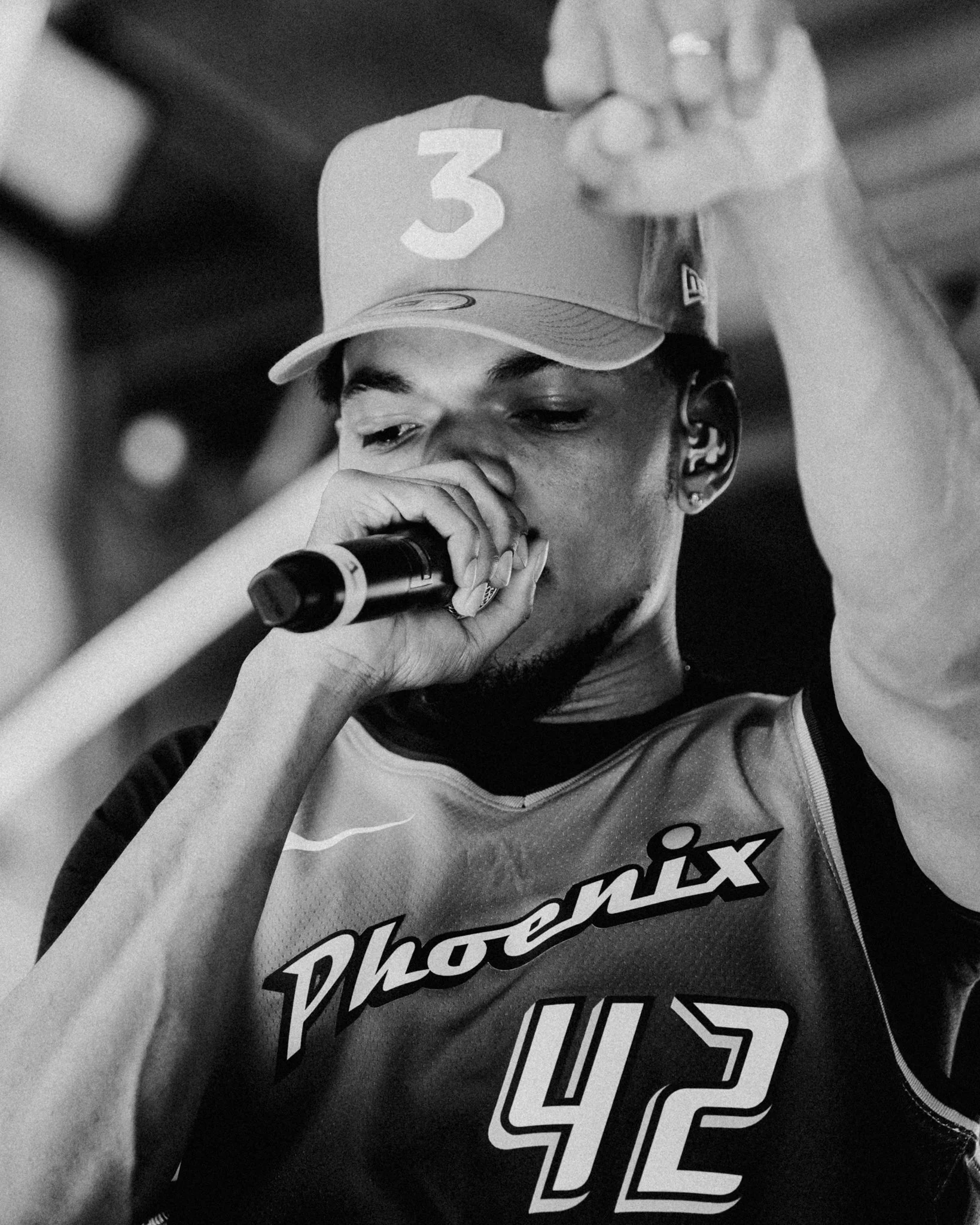 Black and white close-up of a young man wearing a cap with the number 3 and holding a microphone near his mouth, with text on his shirt reading 'Photos' and other partially visible words.