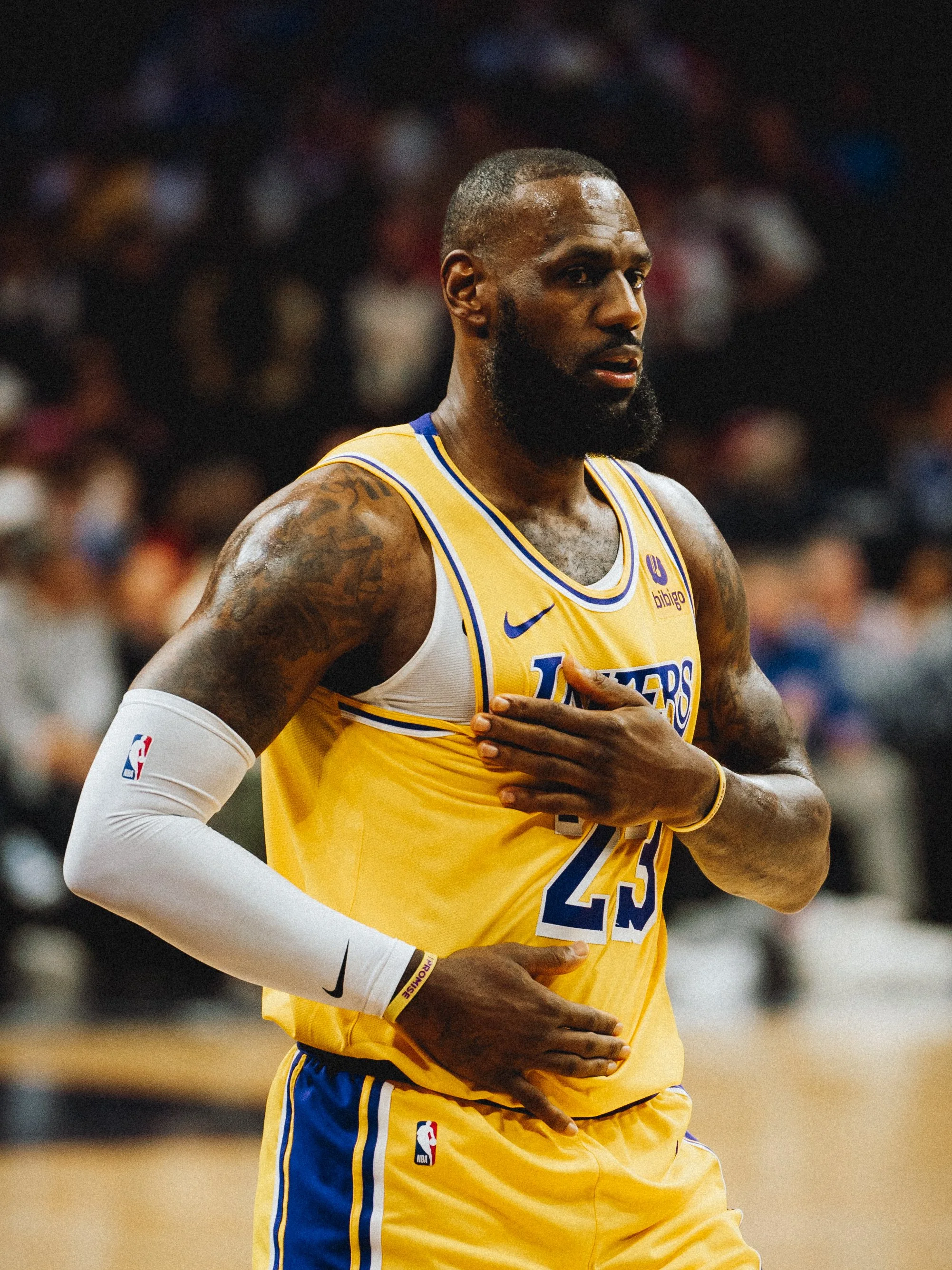 NBA Player, LeBron James from the Los Angeles Lakers, wearing a yellow jersey and white sleeve, standing on a basketball court with a blurred crowd in the background.