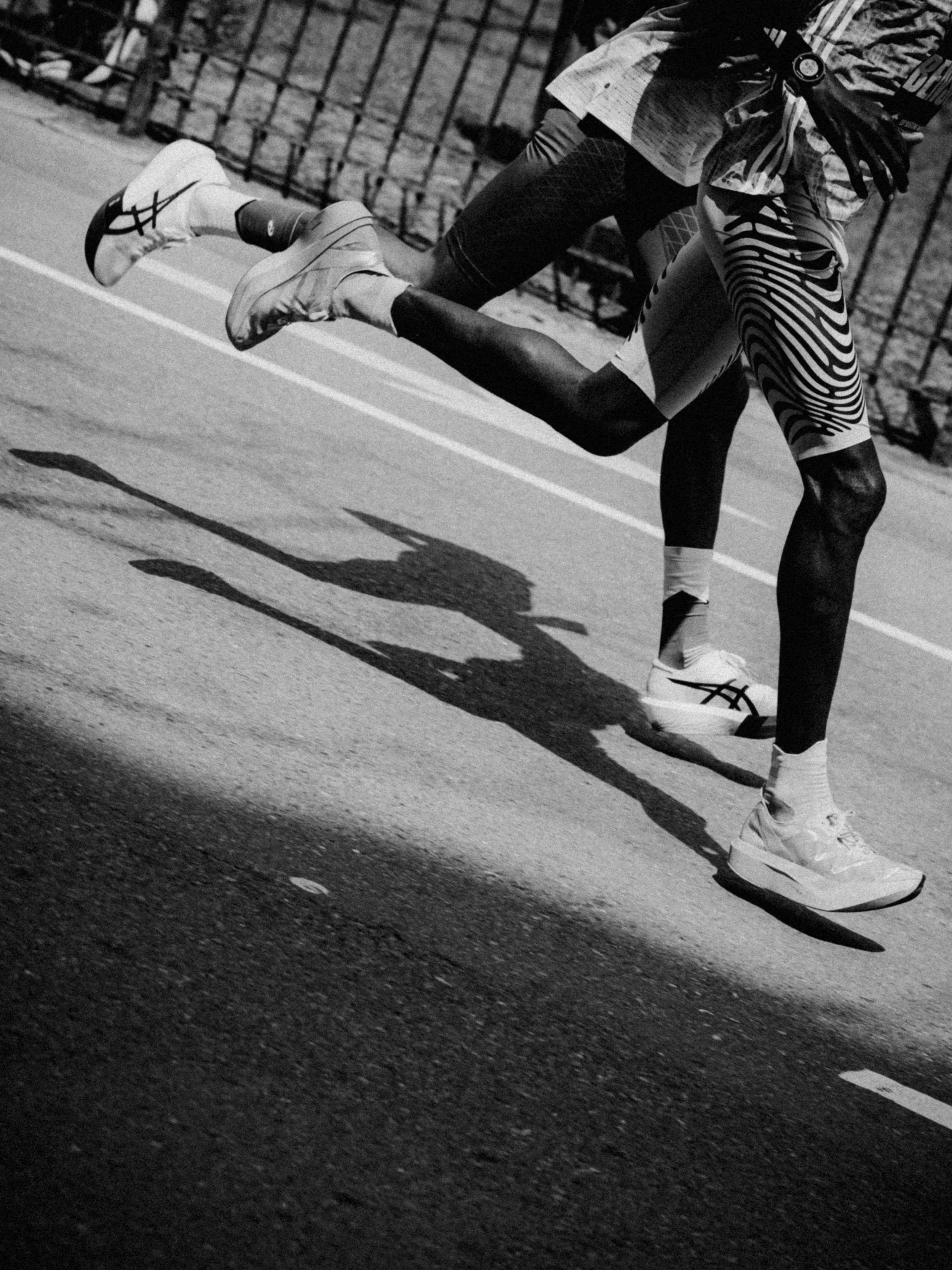 Black and white photo of a runner leaning forward against a chain-link fence, wearing sneakers and sportswear, casting a long shadow on the pavement.