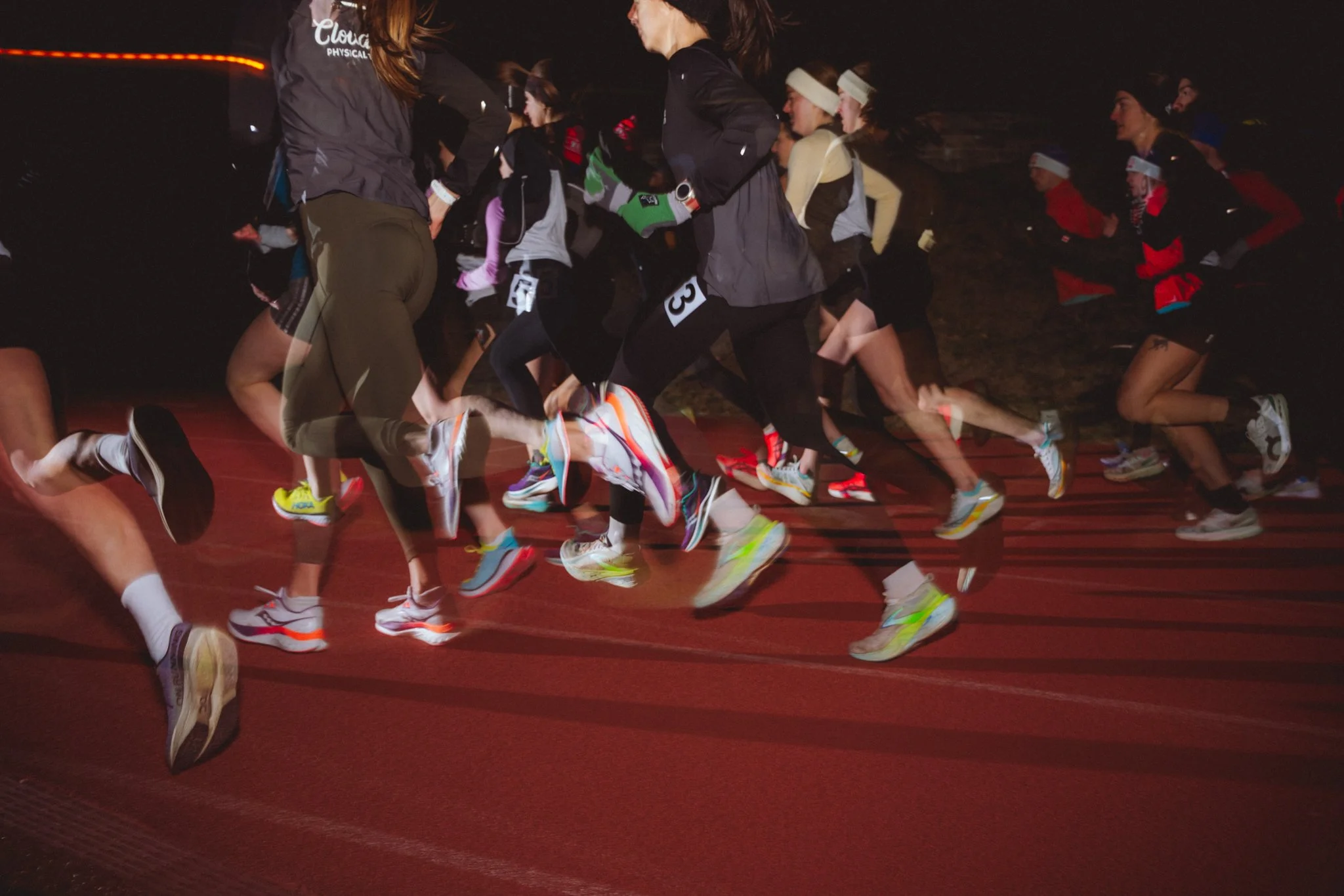Nighttime indoor track race showing a group of runners wearing athletic gear and colorful running shoes, sprinting on a red track with some people wearing headbands and jackets.