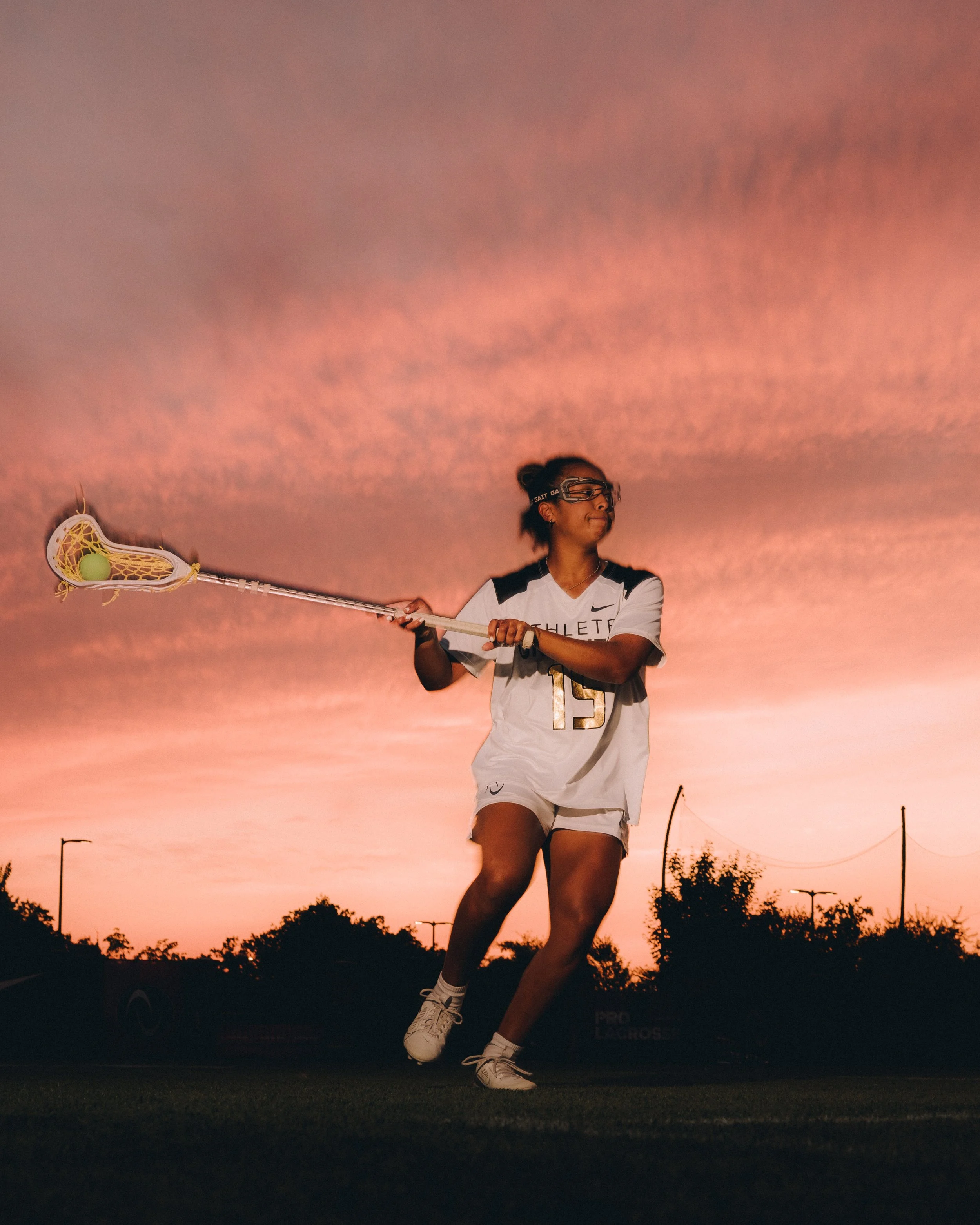 A female lacrosse player stands on the field against a sunset sky, holding a lacrosse stick with a ball in the net. She wears a white and black sports uniform, goggles, and white sneakers. The background shows trees, streetlights, and power lines.