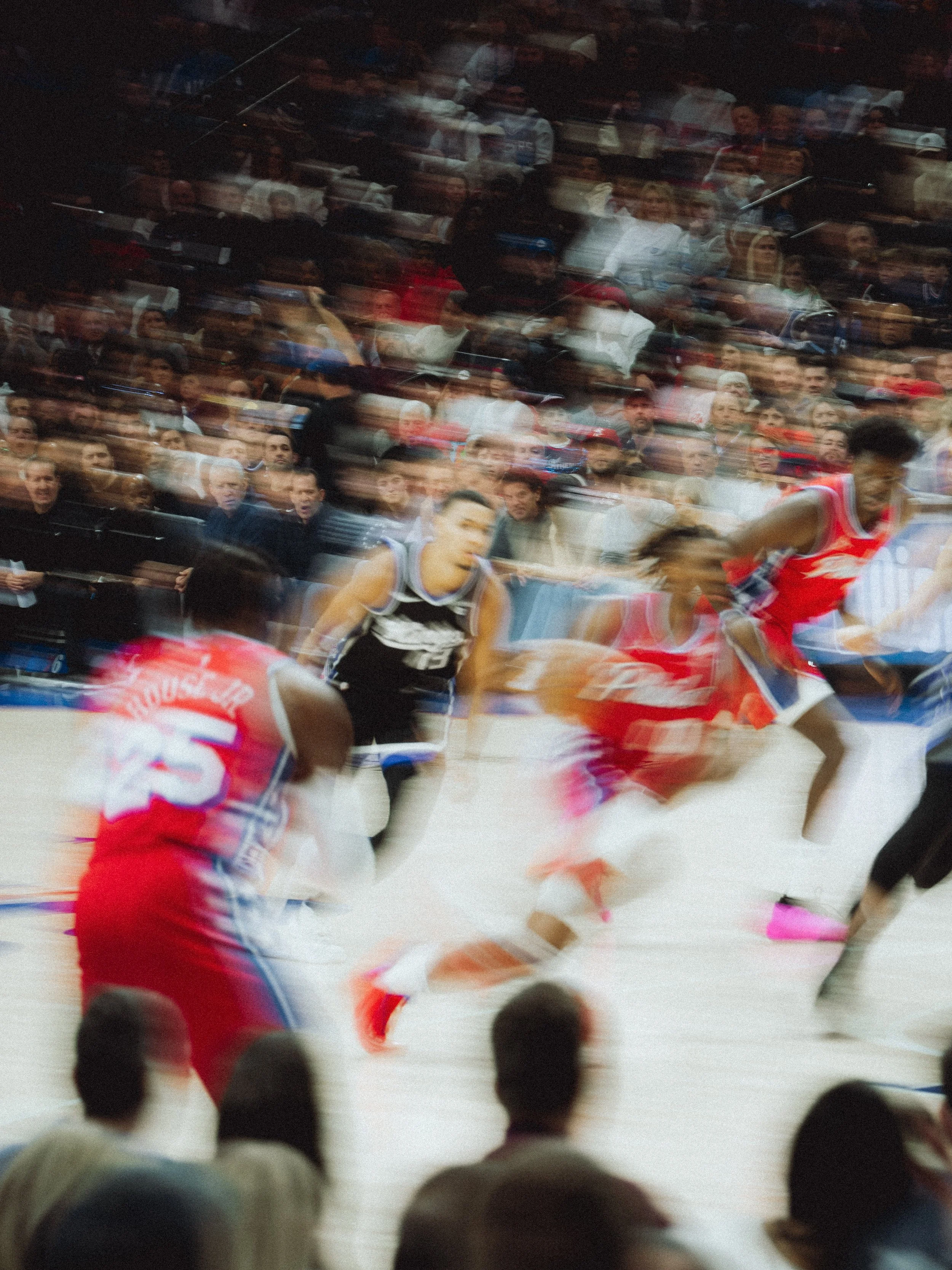 A basketball game is happening with players in red and black jerseys, and spectators watching from the stands. The motion blur indicates fast movement.