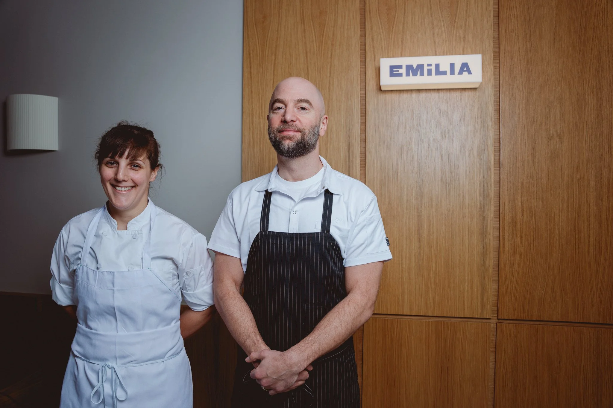 Two chefs, a woman with brown hair and a man with a beard, standing in front of a wooden wall with a sign that reads 'EMILIA' in blue letters.