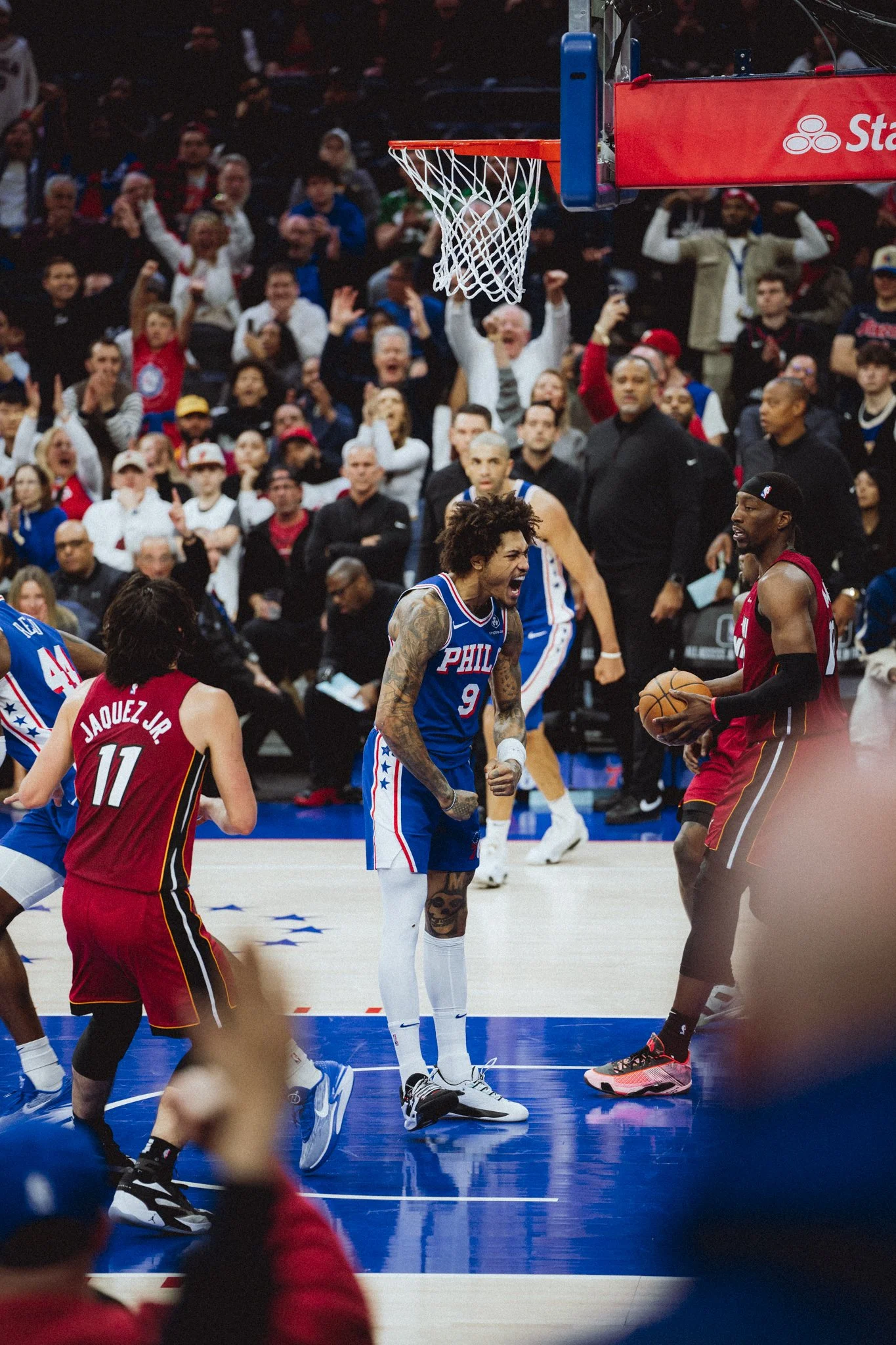 A basketball game with players from the Philadelphia 76ers and Miami Heat on the court. The crowd is cheering, and one player from the Miami Heat is holding the basketball while others are reacting.
