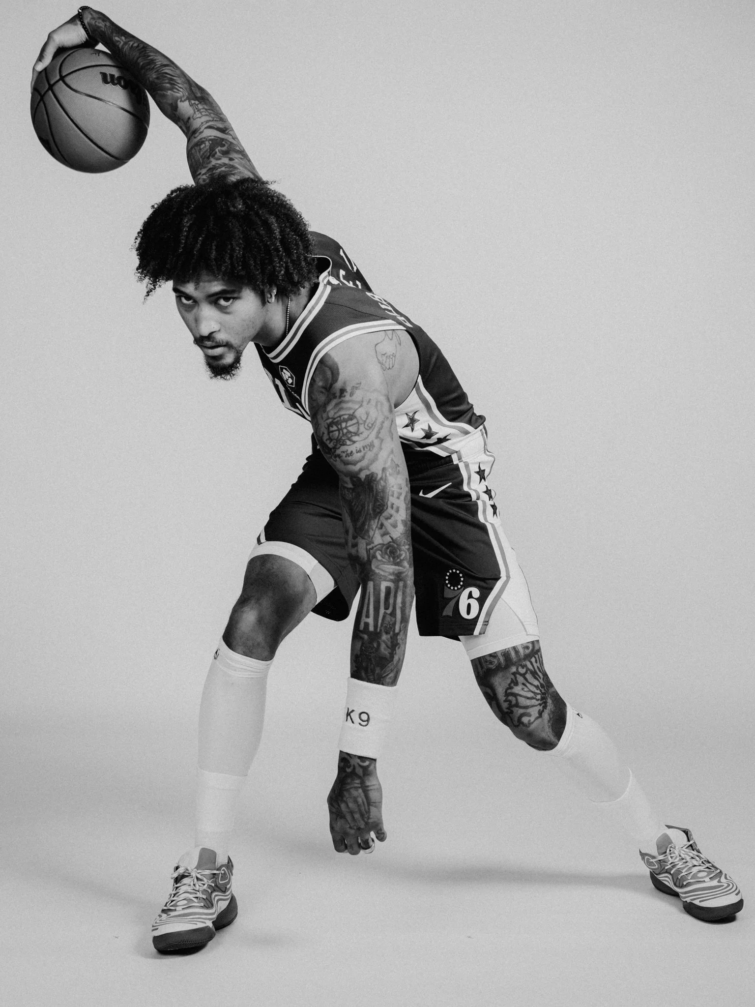 NBA Player,  Kelly Oubre in athletic clothing bending down, holding a basketball with his right hand, with tattoos on his arms and legs, wearing sneakers, and looking at the camera in a studio with a plain background.