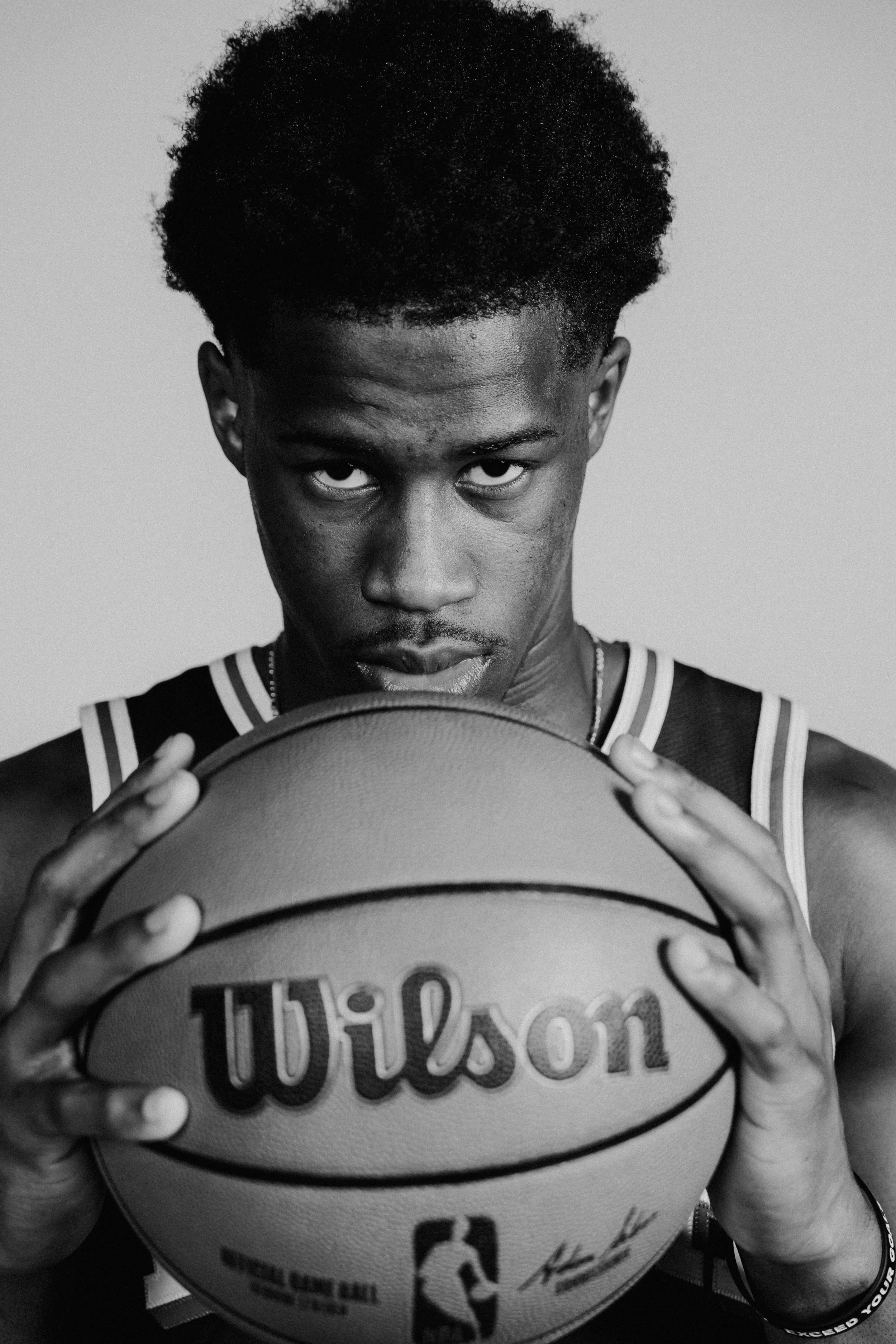 NBA Player,  VJ Edgecombe with short curly hair and a serious expression holds a basketball close to his face, looking directly at the camera. He is wearing a sports jersey and a wristband.