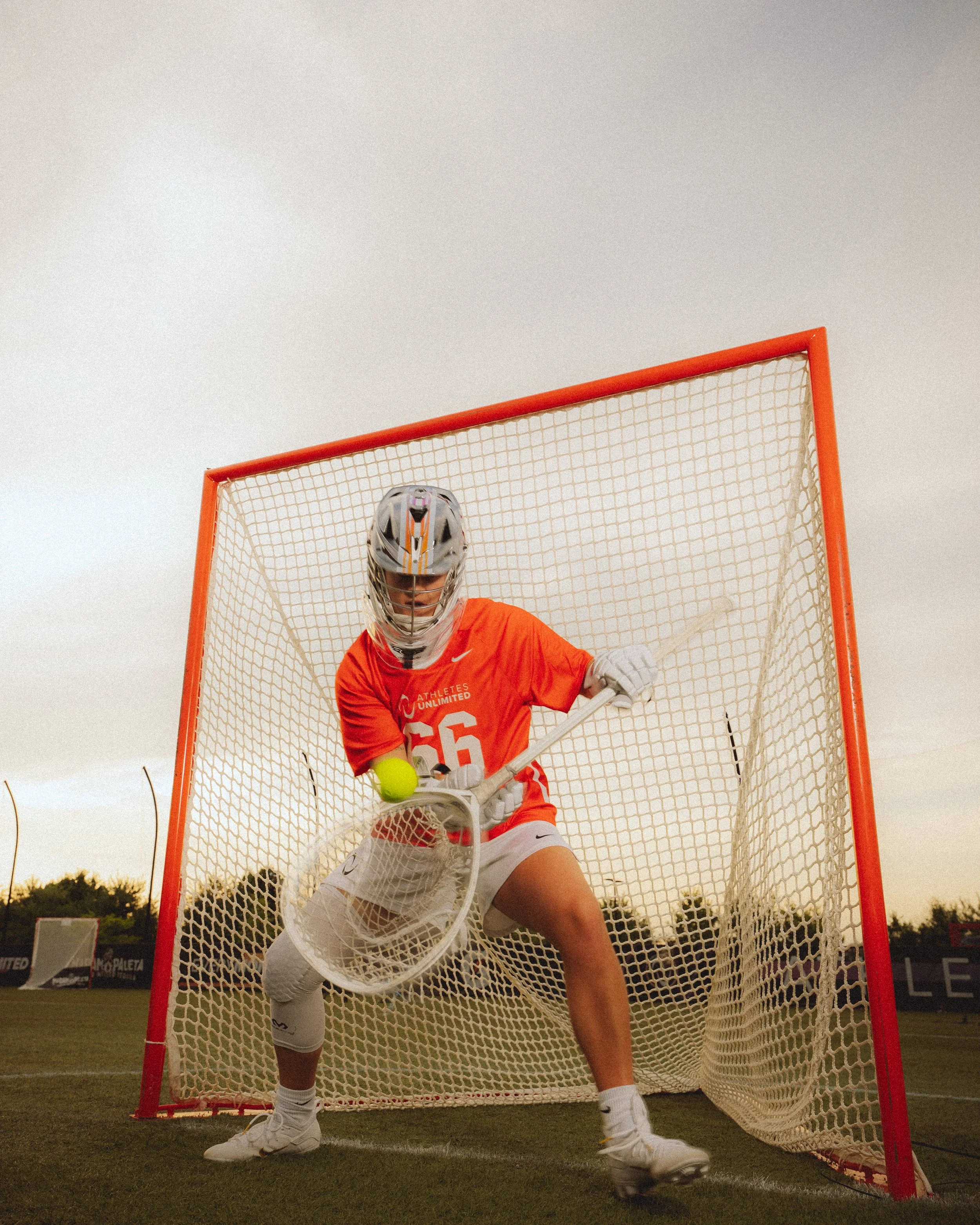 Hockey goalie in orange jersey and white pads crouches in front of net, preparing to stop a yellow puck, during sunset on a grassy field.