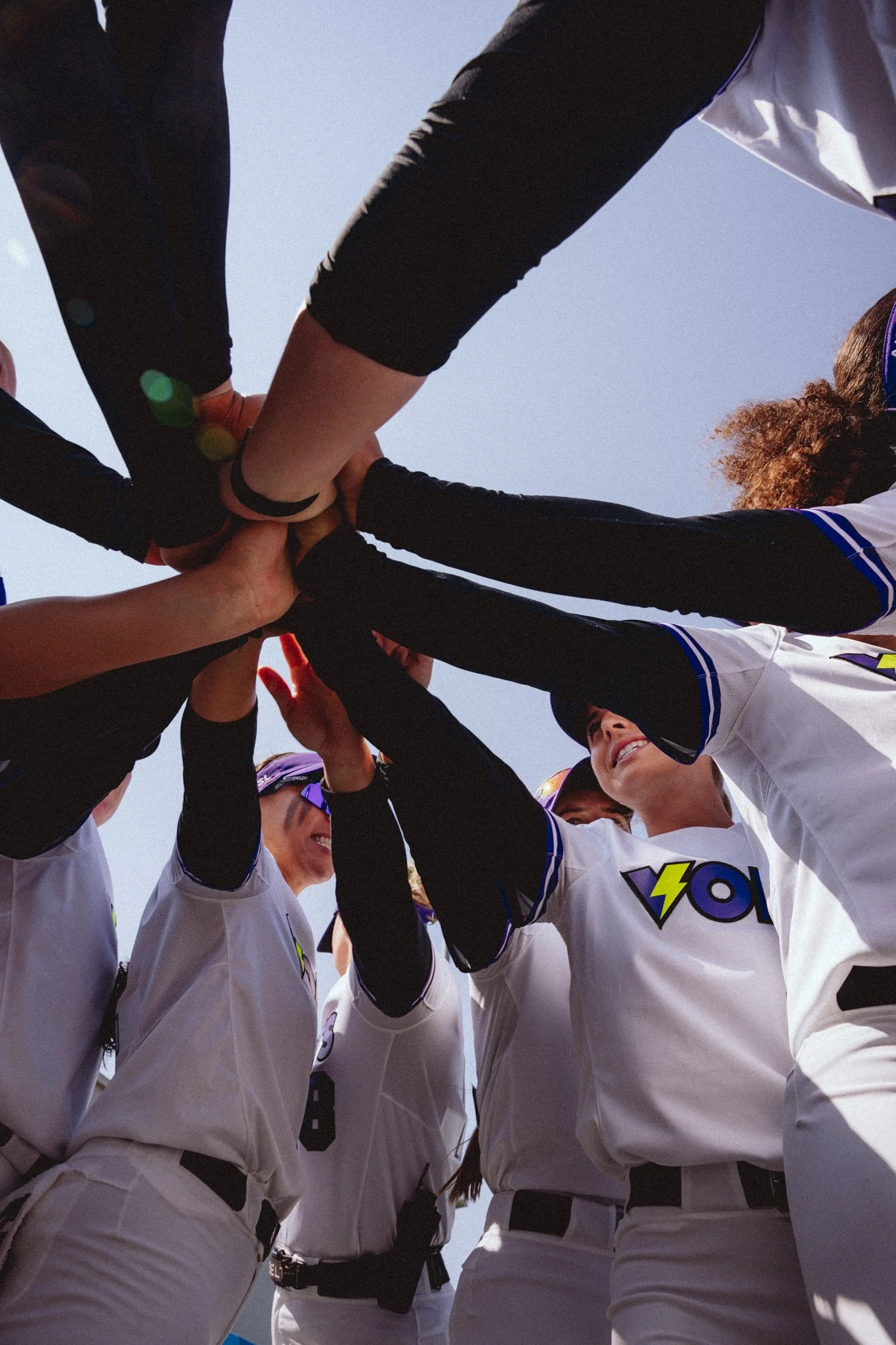 A group of youth athletes dressed in white and black sports uniforms participating in a team huddle with hands stacked together in the center, smiling and looking up at each other against a bright blue sky.