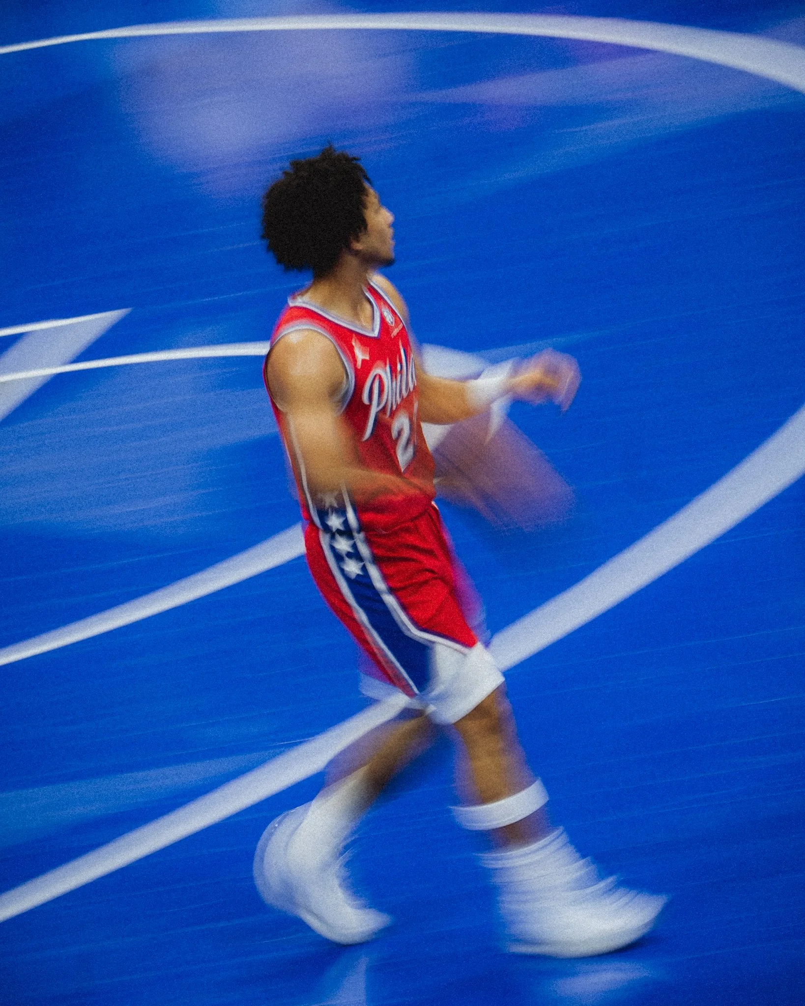 A basketball player in a red Philadelphia 76ers jersey with the number 2 on it, dribbling the ball on a blue basketball court with white lines, captured with motion blur.