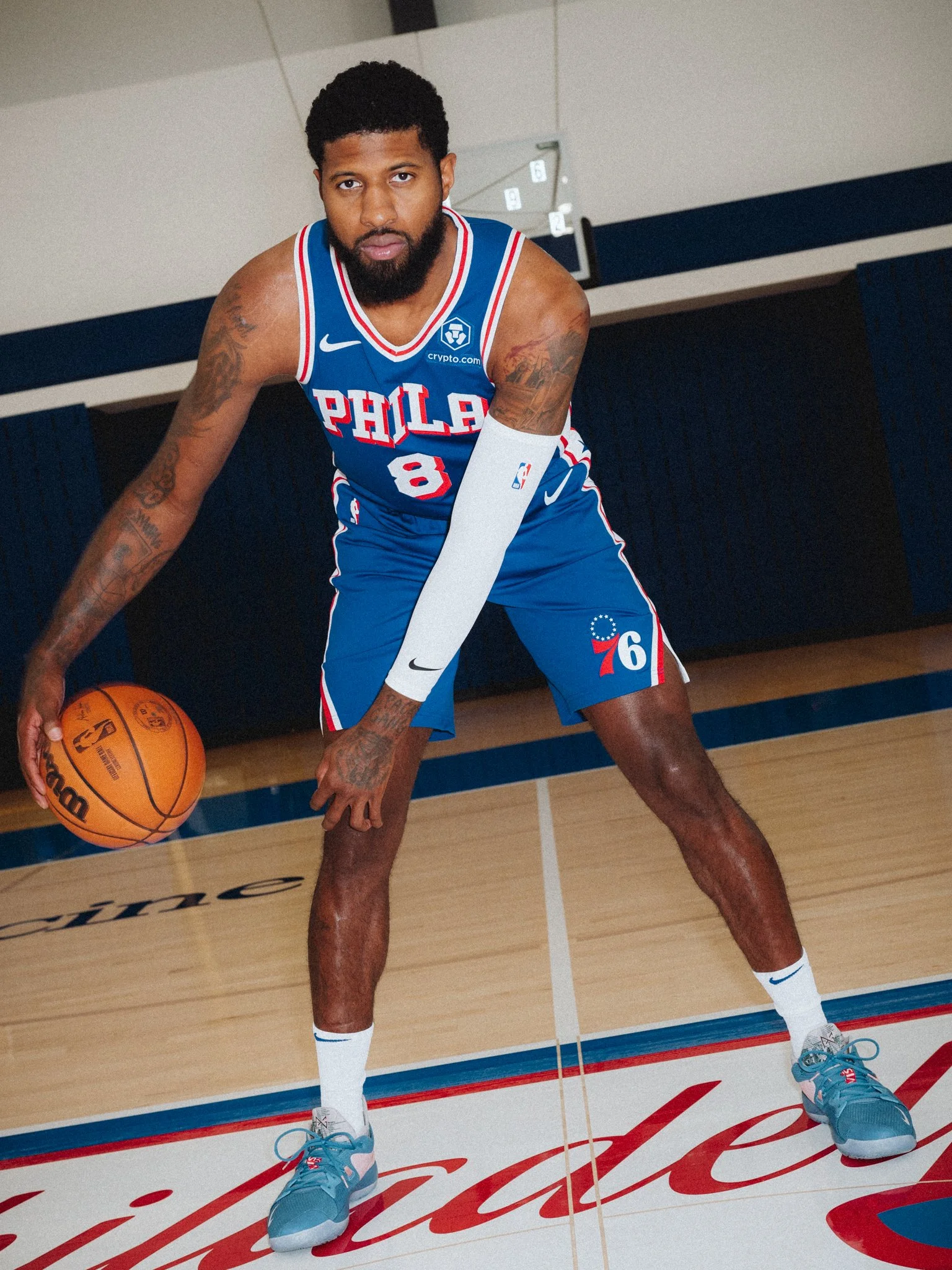 A male basketball player in a blue Philadelphia 76ers uniform, wearing blue sneakers, is playing on a basketball court. He is holding a basketball with his left hand.