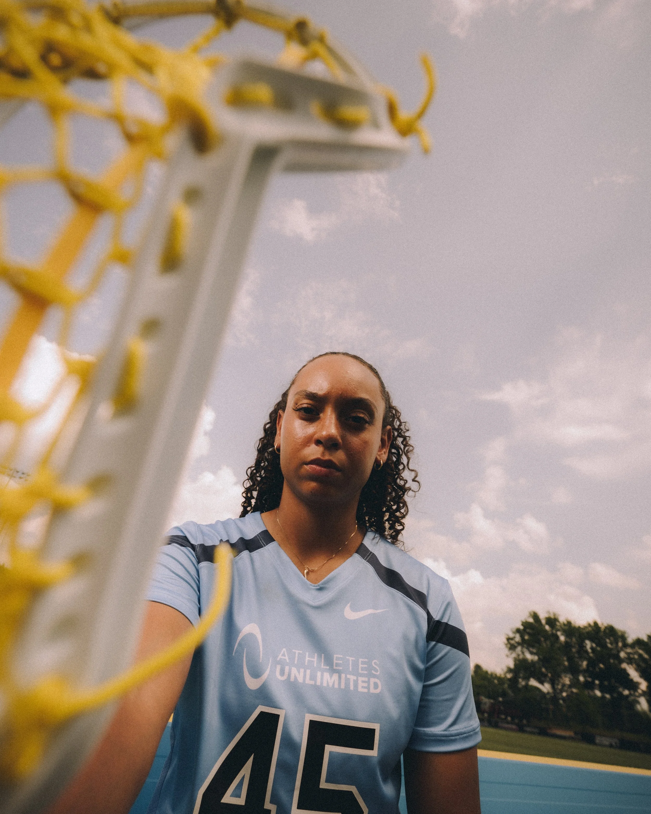 A female athlete with curly hair taking a selfie on a sports field, with a focus on her face and a soccer goal in the foreground, during daylight with a cloudy sky.