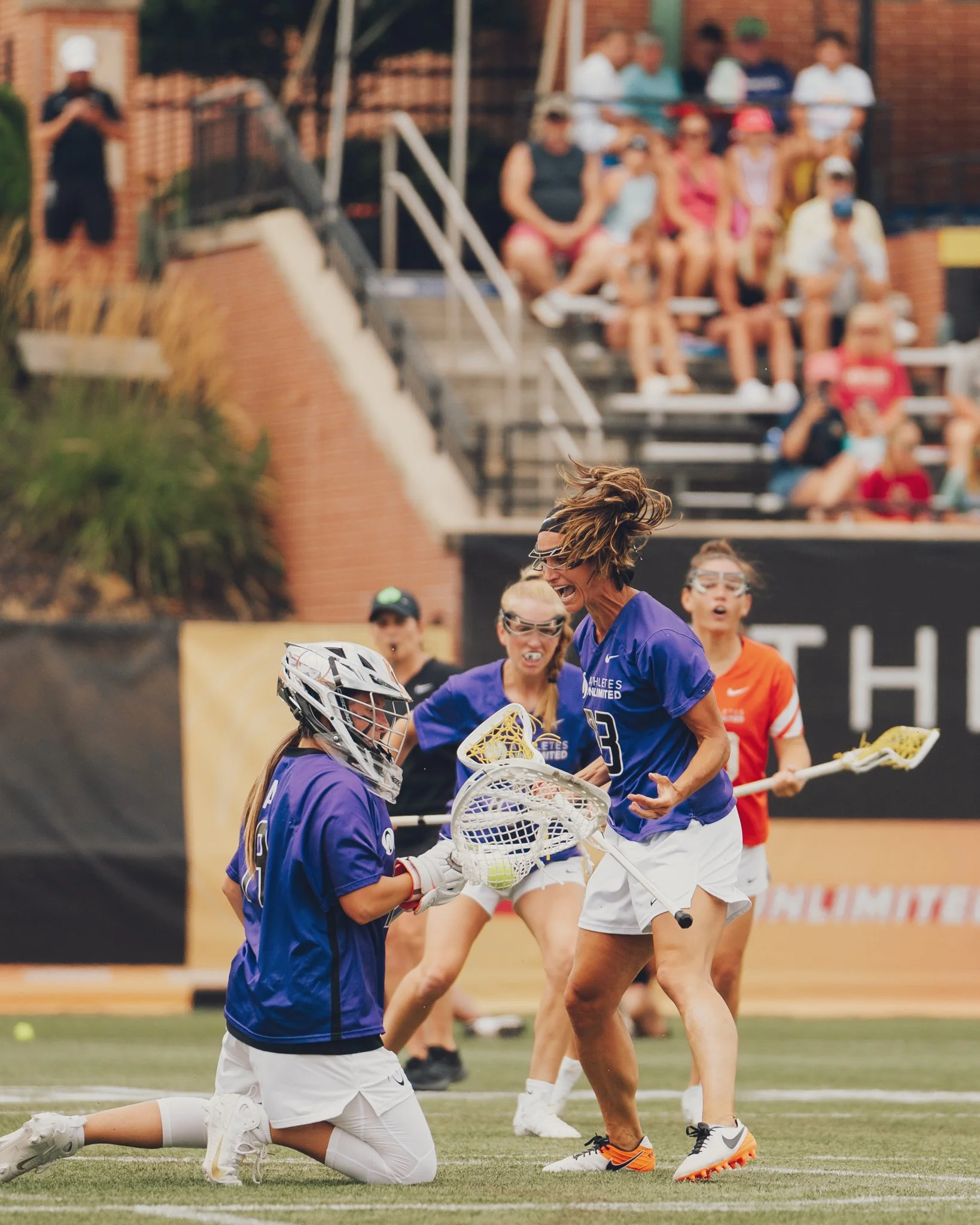 Lacrosse players in action during a game, with a player in a purple jersey and another in black gear, including a helmet and gloves, on a grassy field. Spectators watch from the bleachers in the background.