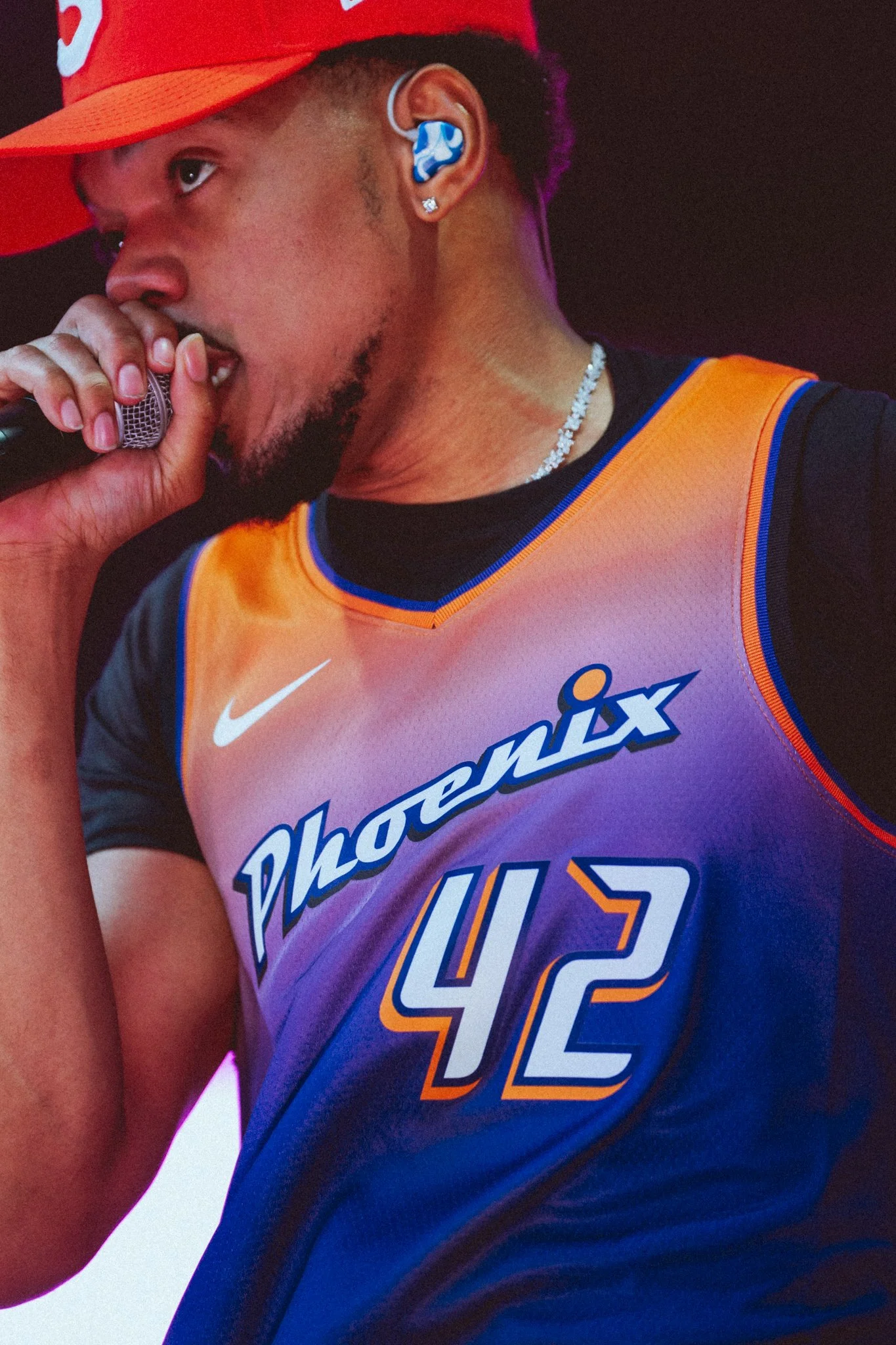 Close-up of a basketball player wearing a Philadelphia 76ers jersey with the number 42, an orange and black uniform, a red cap, and earrings, holding a microphone near his mouth.