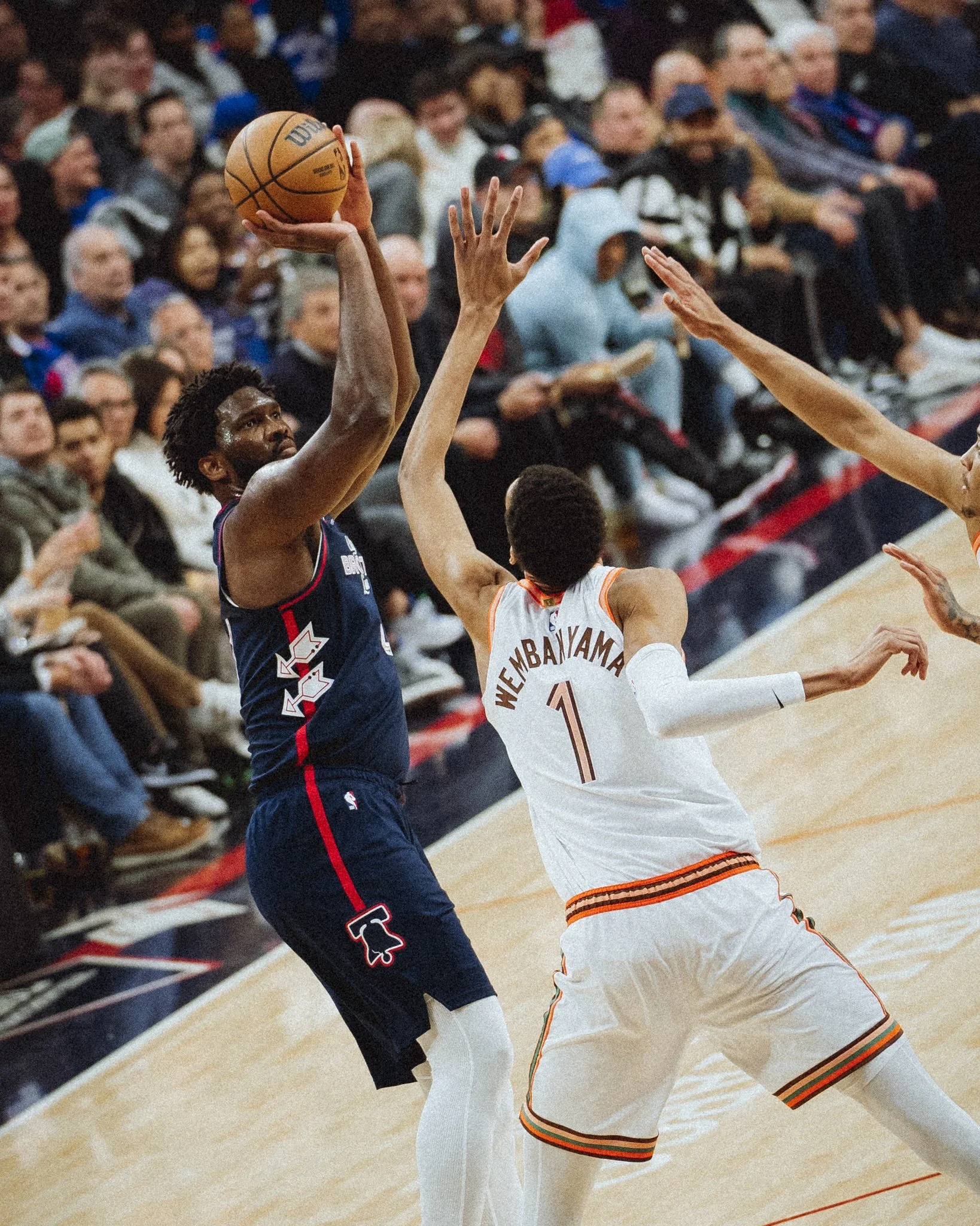 Two basketball players in action during a game, one wearing a navy blue jersey with red and white accents holding the ball, and the other wearing a white jersey with orange and black trim defending. Spectators seated in the background.