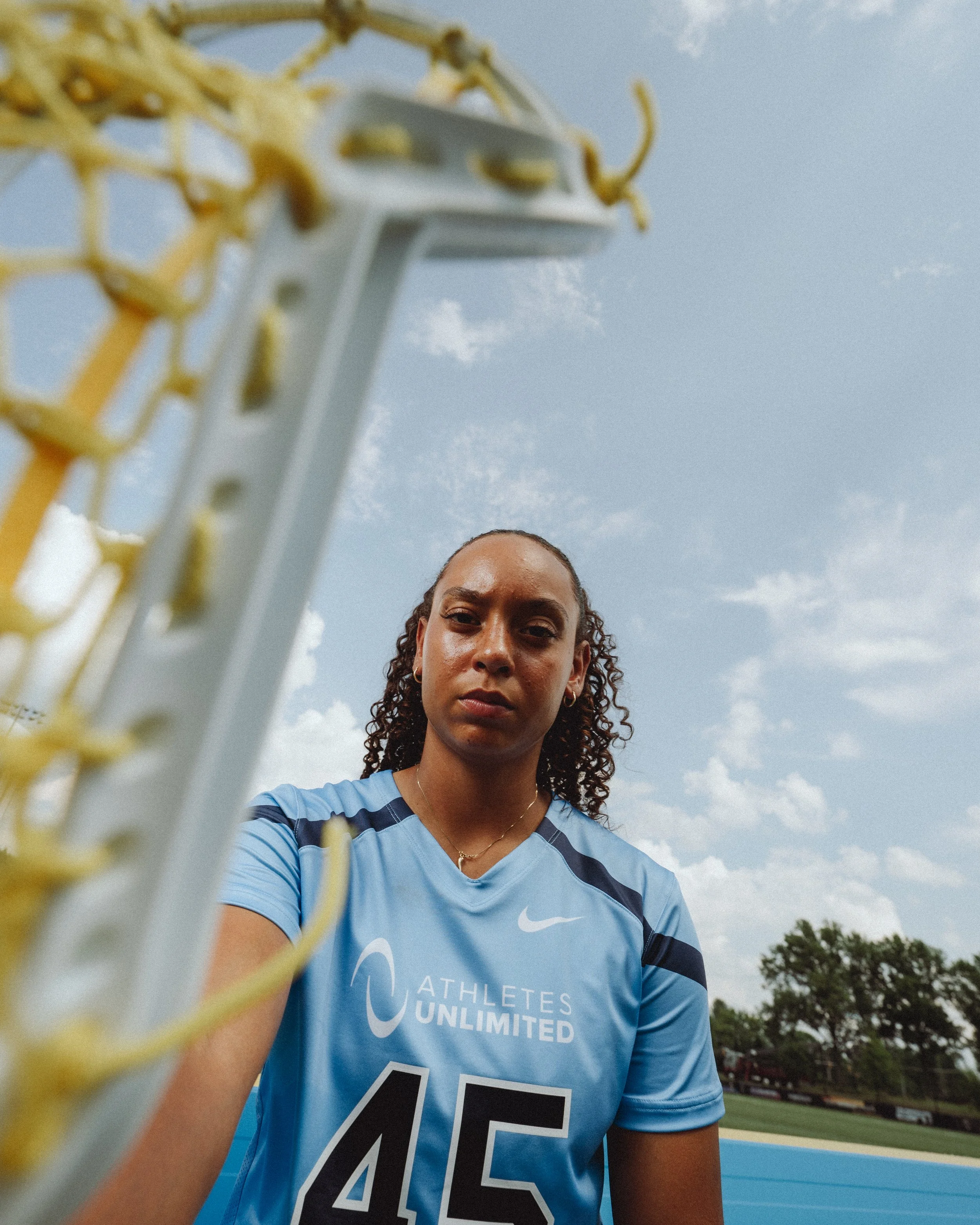 A female athlete wearing a blue sports jersey with the text 'ATHLETES UNLIMITED' and the number 45 on it, standing outdoors on a cloudy day near a sports field, with a chain-link fence and trees in the background, as seen from a low angle.