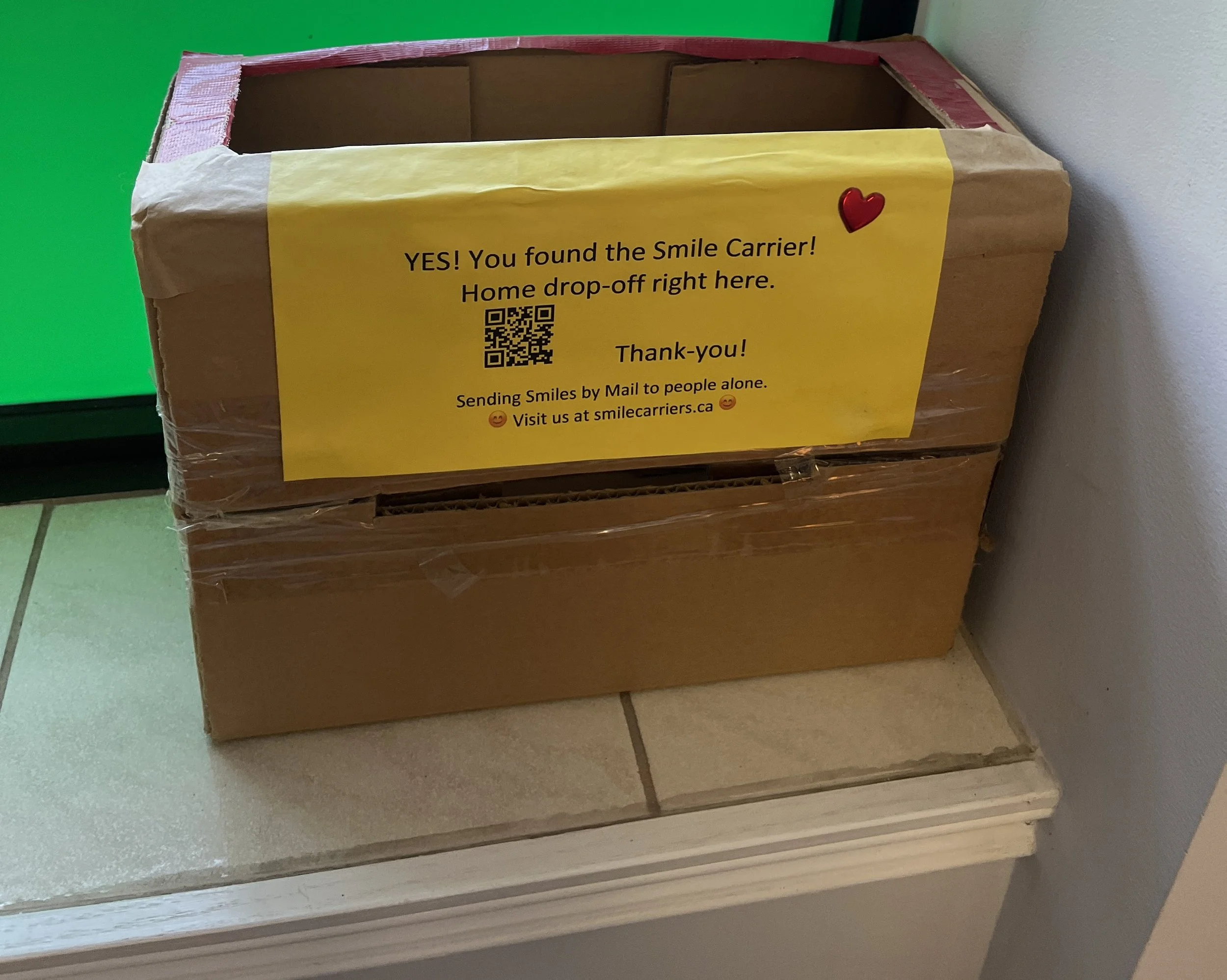 A cardboard box with a yellow sign attached to it, indicating it is a Smile Carrier for home drop-off. The sign includes a QR code and a message about sending smiles by mail.