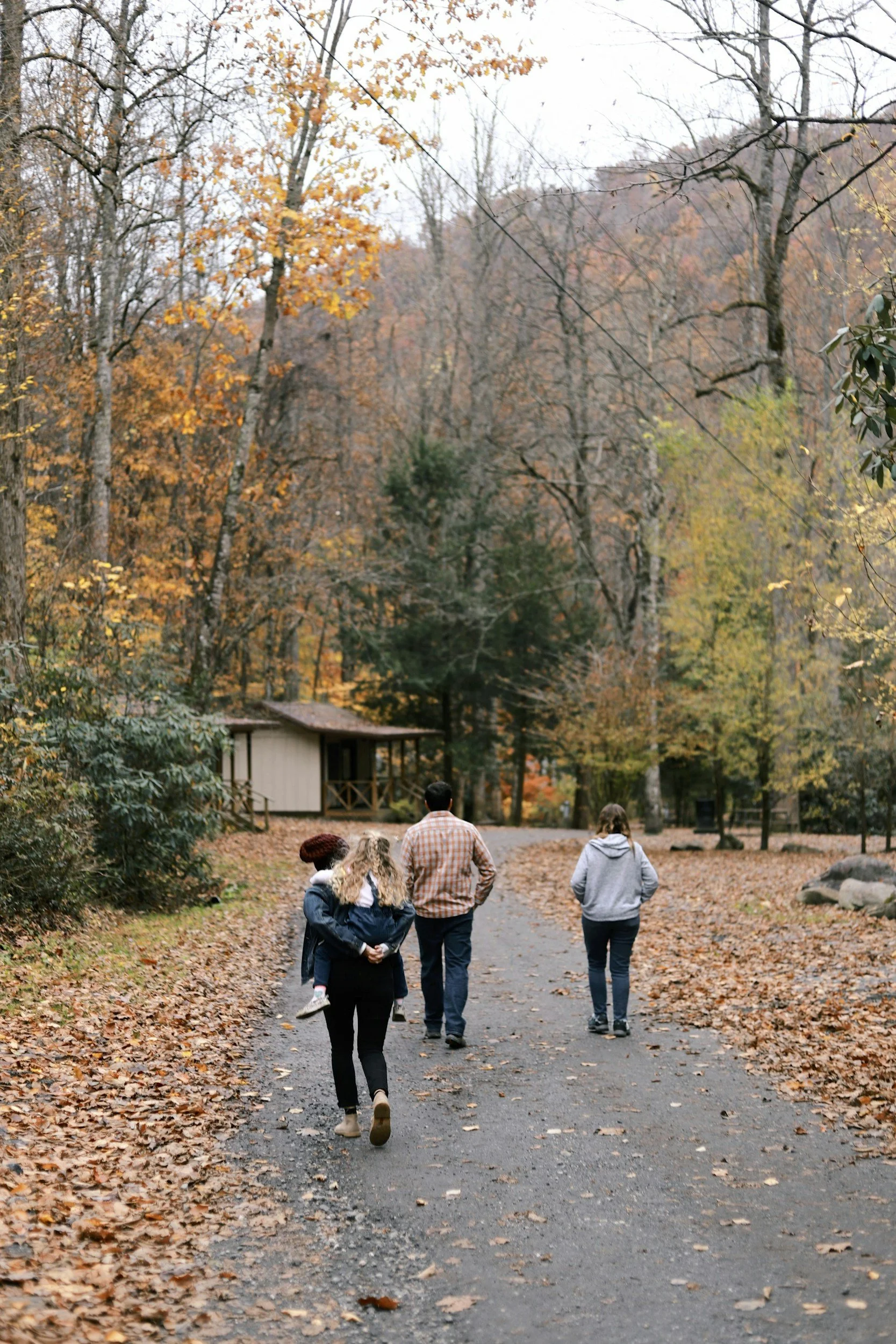 People walking on a leaf-covered path in a wooded area during fall, with trees showing autumn colors and a small building visible in the background.