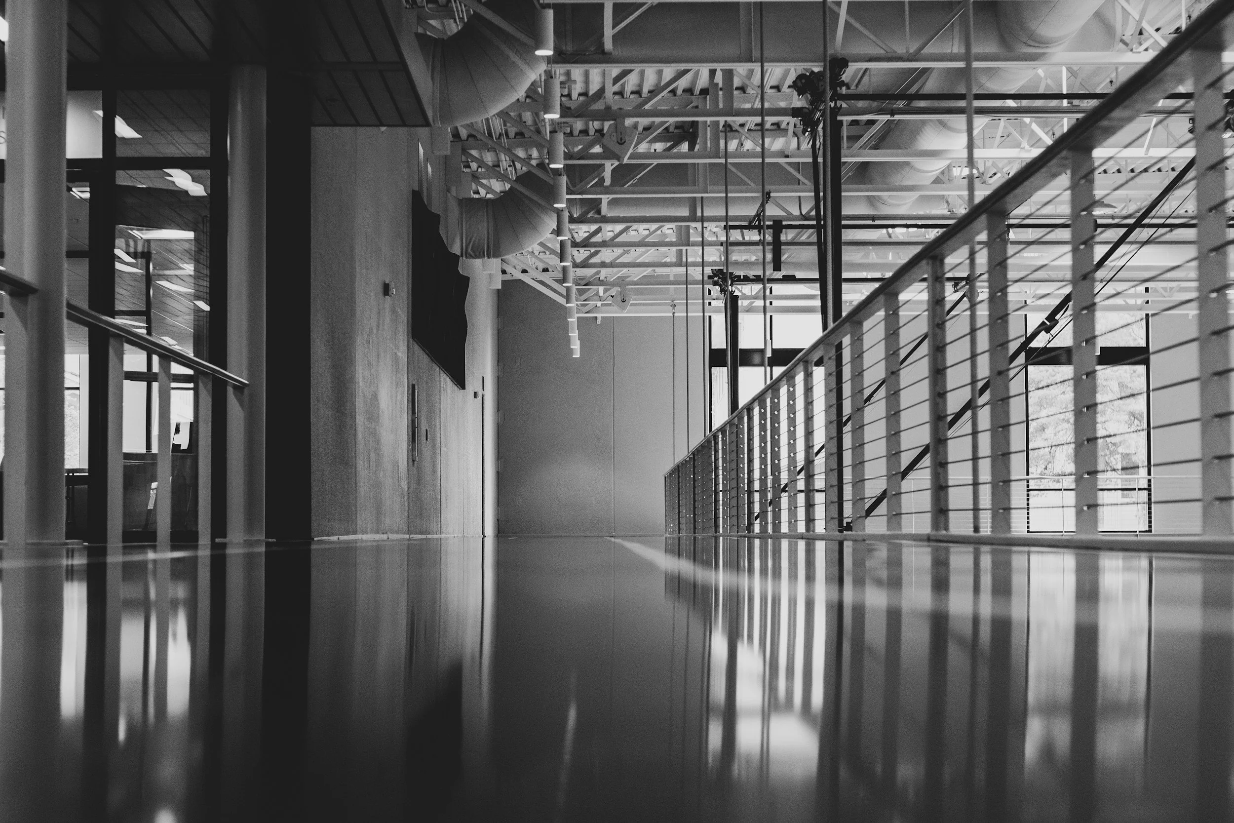 Black and white interior view of a modern building with a corridor, metal railing, large windows, visible ceiling pipes and structures, and a wooden floor.