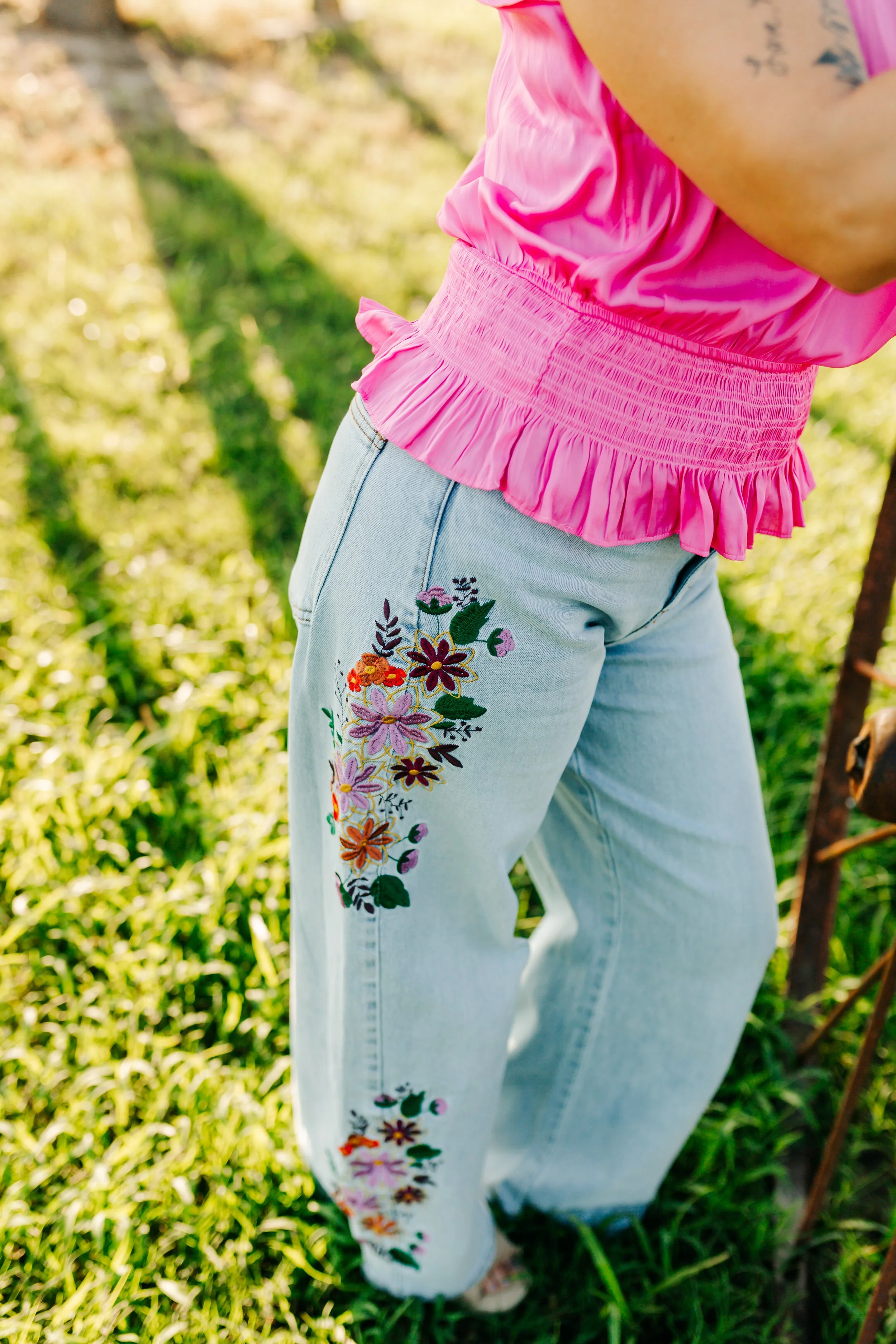 Close-up of a person wearing light blue jeans with colorful floral embroidery on the side and a pink ruffled top, standing outdoors on green grass.