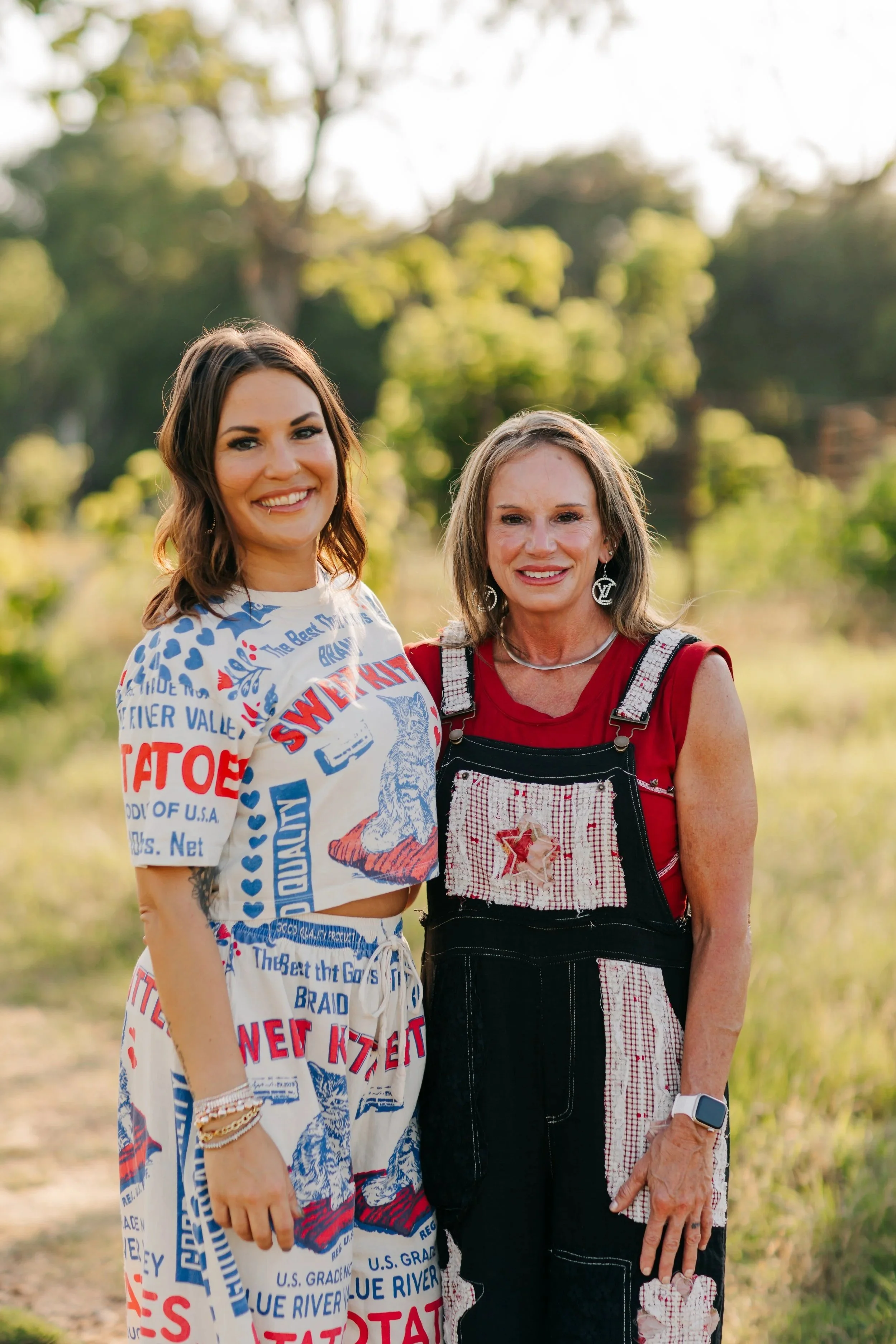 Two women standing outdoors in a sunny setting with greenery in the background, smiling at the camera.