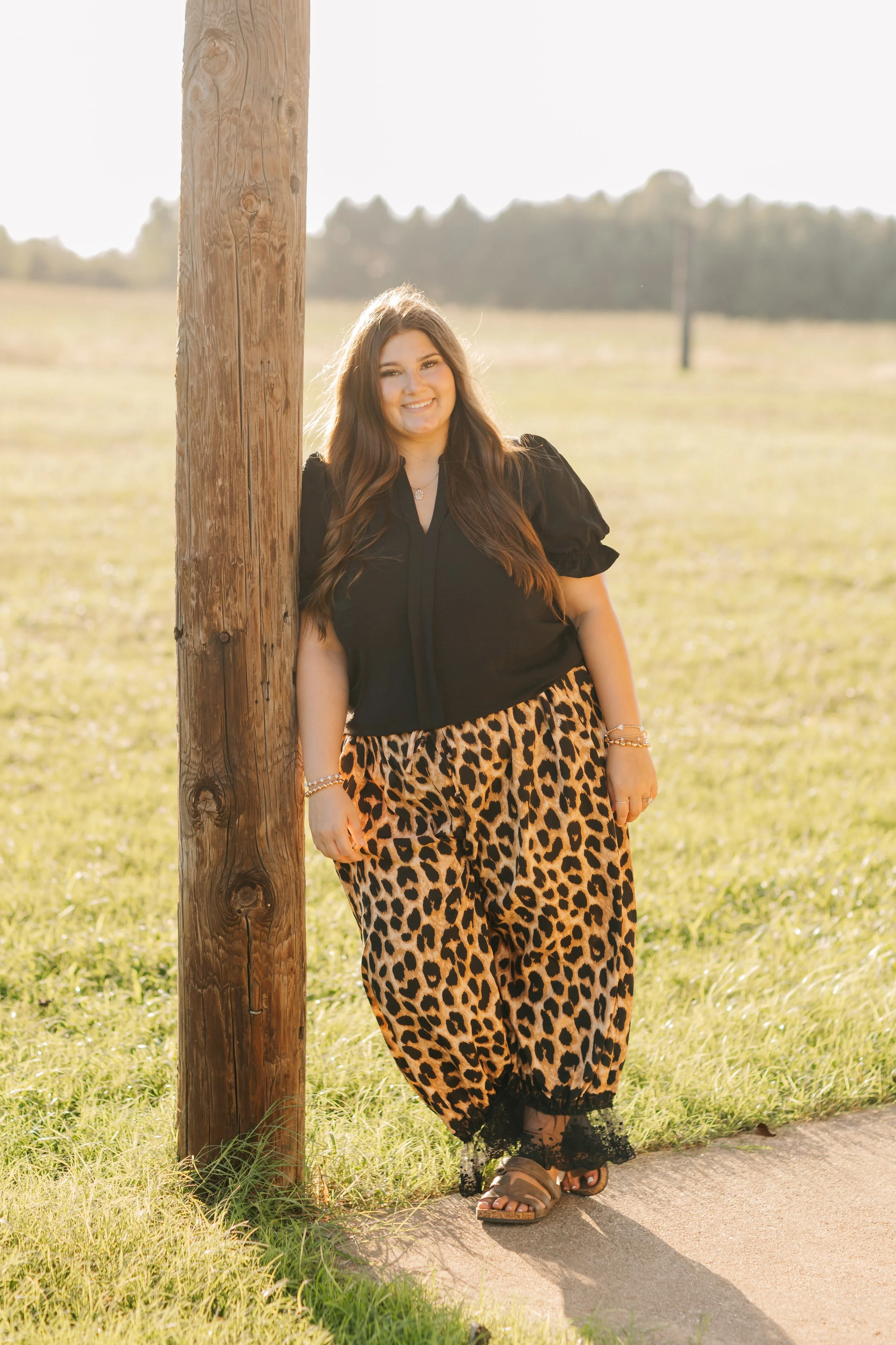 A woman with long brown hair smiling, standing next to a wooden utility pole outdoors on a sunny day, wearing a black top, leopard print pants, and sandals.