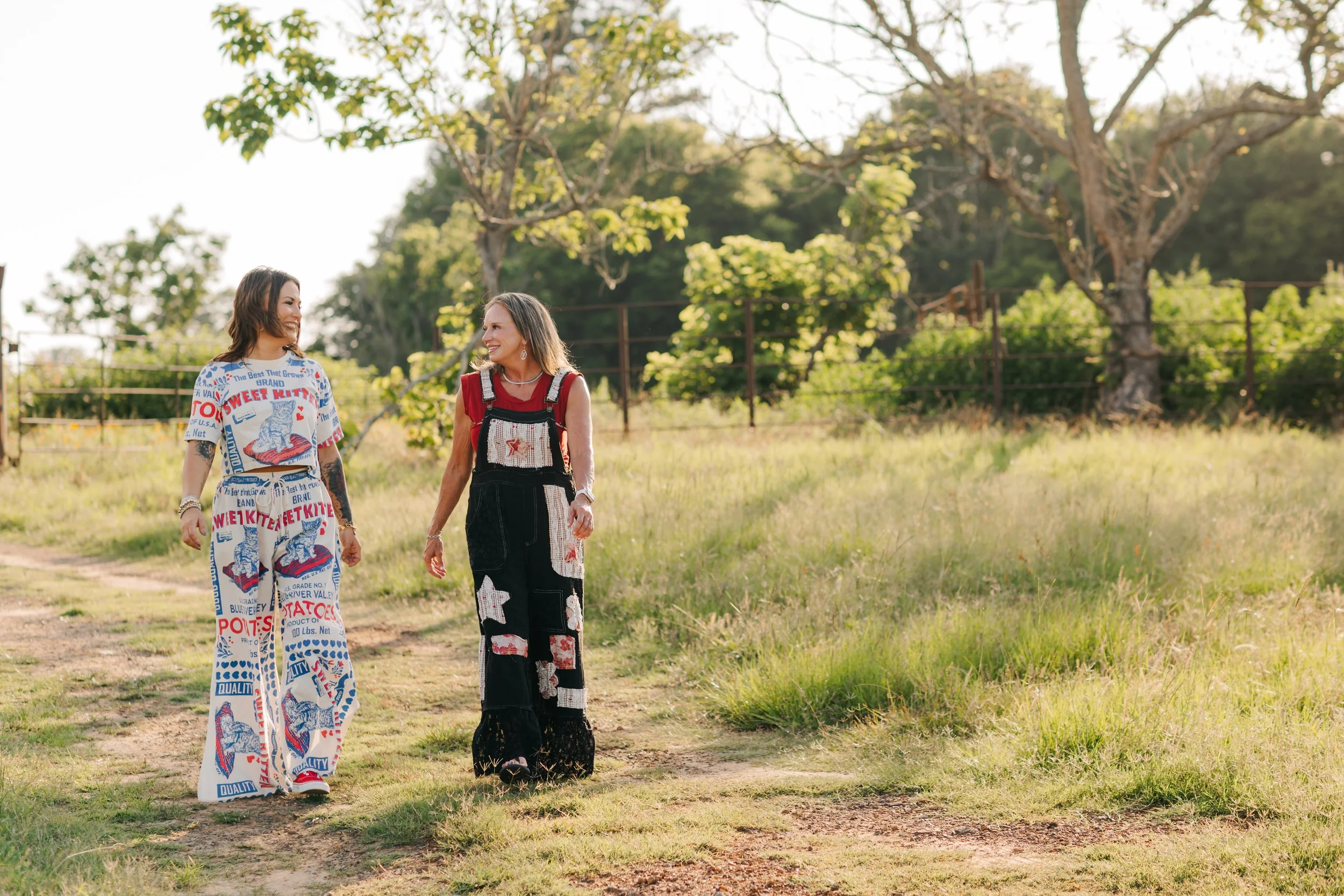 Two women walking outdoors on a grassy path, smiling and chatting, surrounded by trees with green leaves in the background.