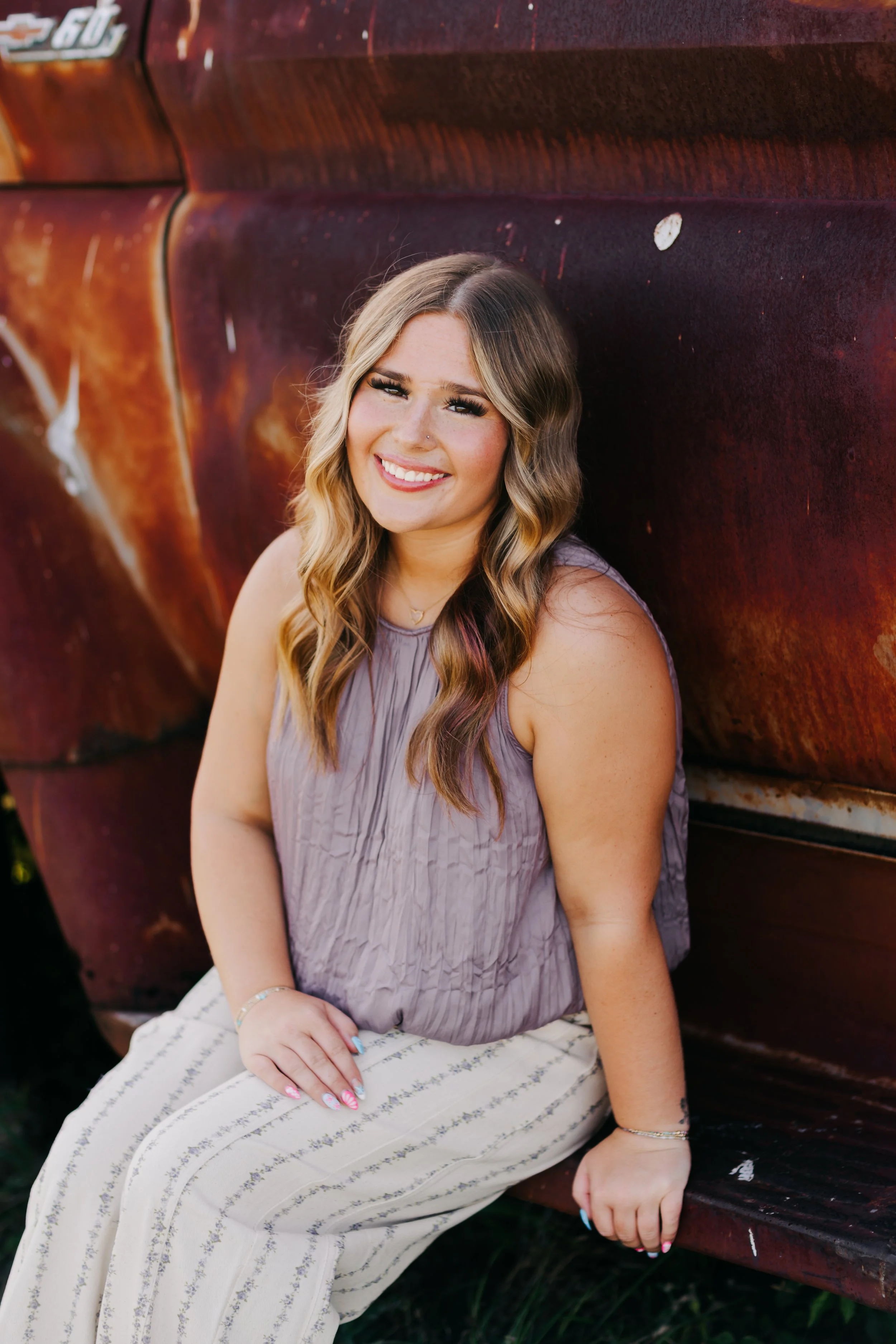 A young woman with wavy, light brown hair sitting on a bench, smiling at the camera, wearing a sleeveless lavender top and striped cream and gray pants.
