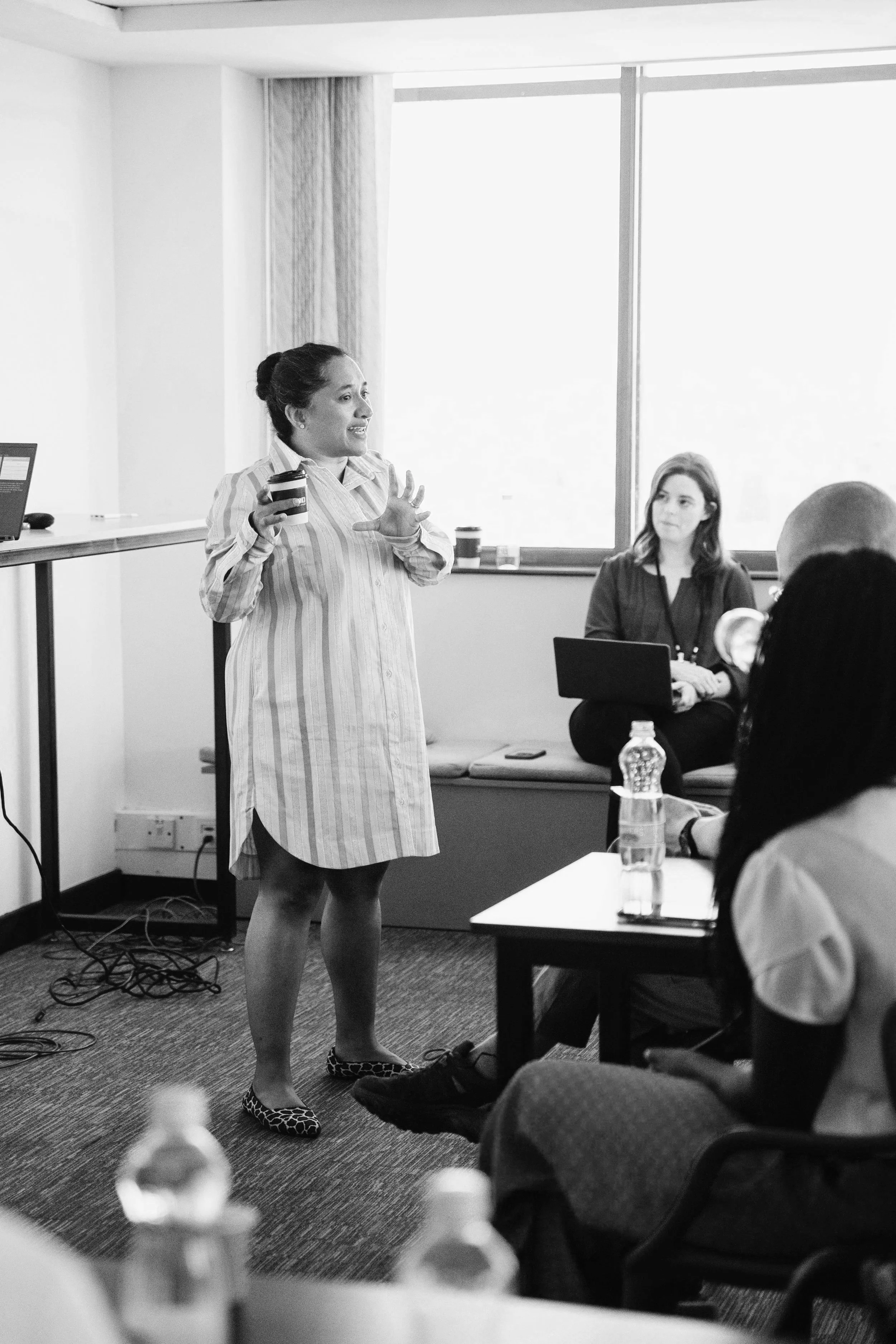 A woman with curly hair tied back, wearing a striped dress and leopard print flats, speaking and gesturing with her hands during a presentation in a conference room with seated audience members.