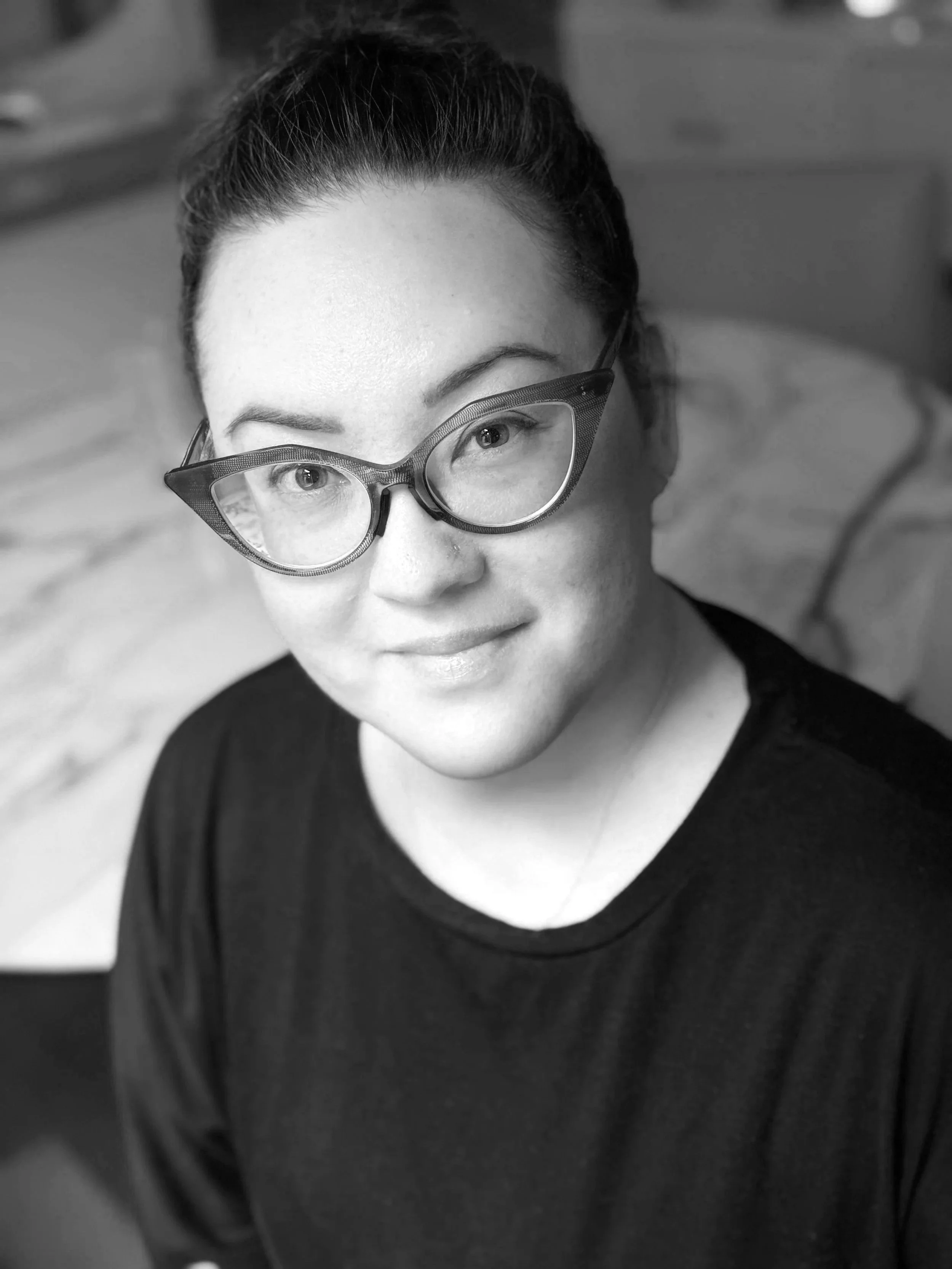 A woman with light skin, dark hair pulled back, and glasses with a thick, textured frame looking at the camera. She is wearing a dark top and smiling slightly.