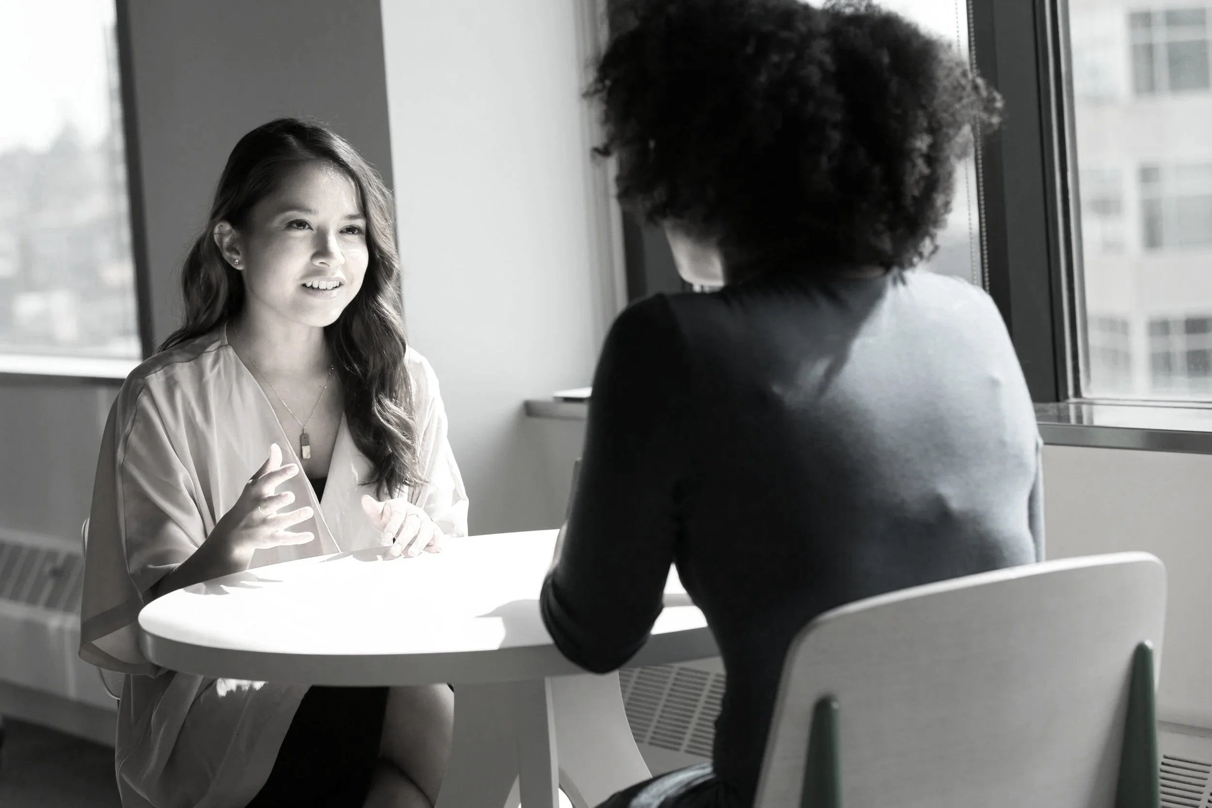 A woman with long wavy hair, wearing a light-colored blouse and necklace, is sitting at a round table and talking to another person with curly hair, who is facing away from the camera near a window in a modern office setting.