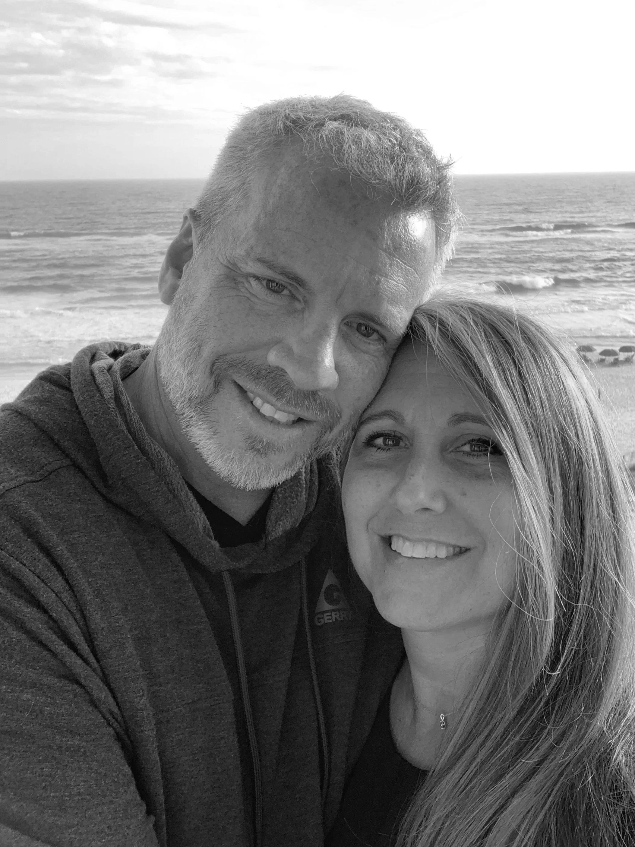 A black and white photo of a man and woman taking a close-up selfie on the beach. The ocean and sky are in the background, and they are smiling with their heads touching.
