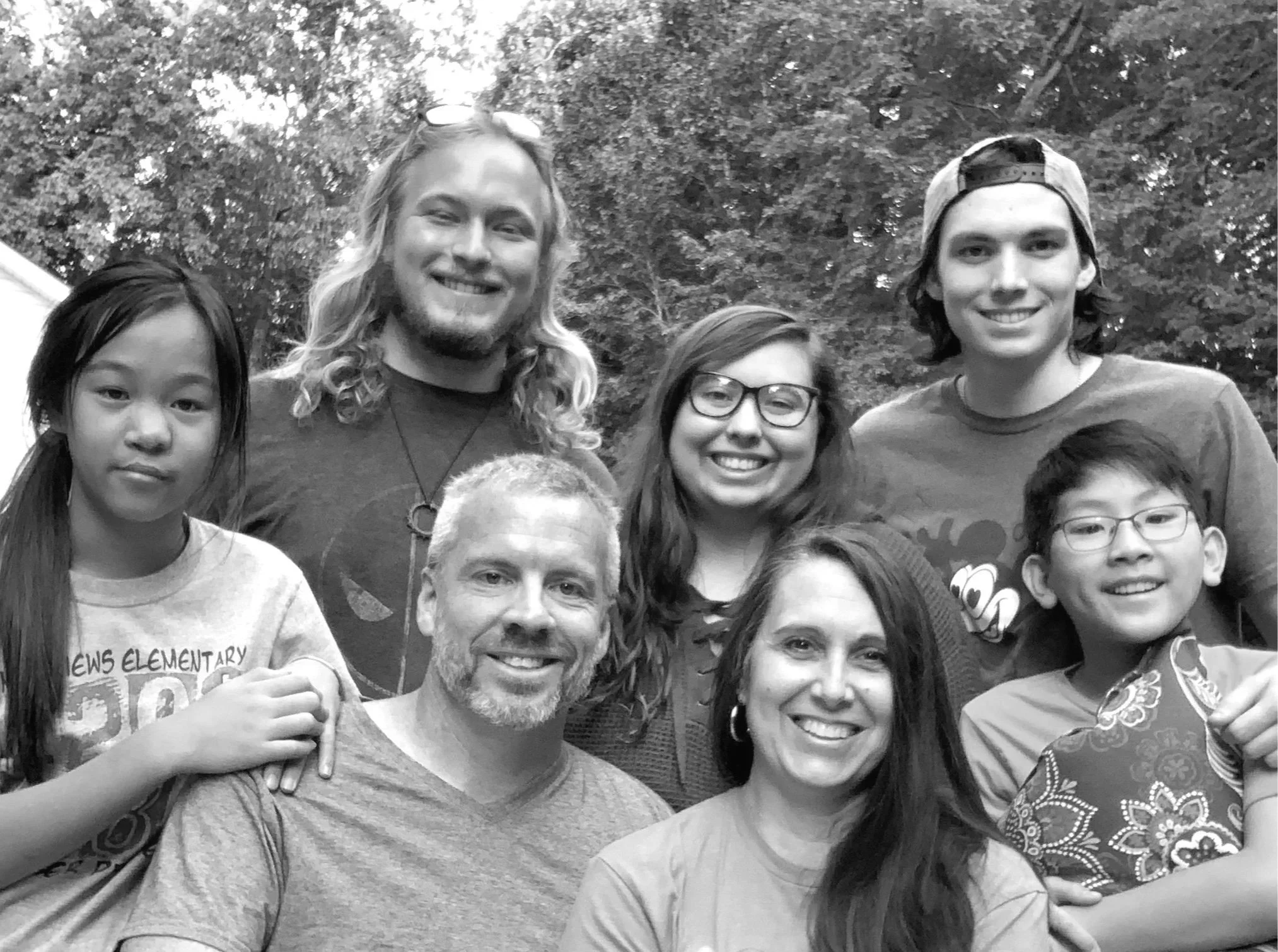 A group of diverse smiling people, including children and adults, gathered outdoors in front of trees, posing for a photo. Some individuals are wearing glasses, hats, and casual clothes.