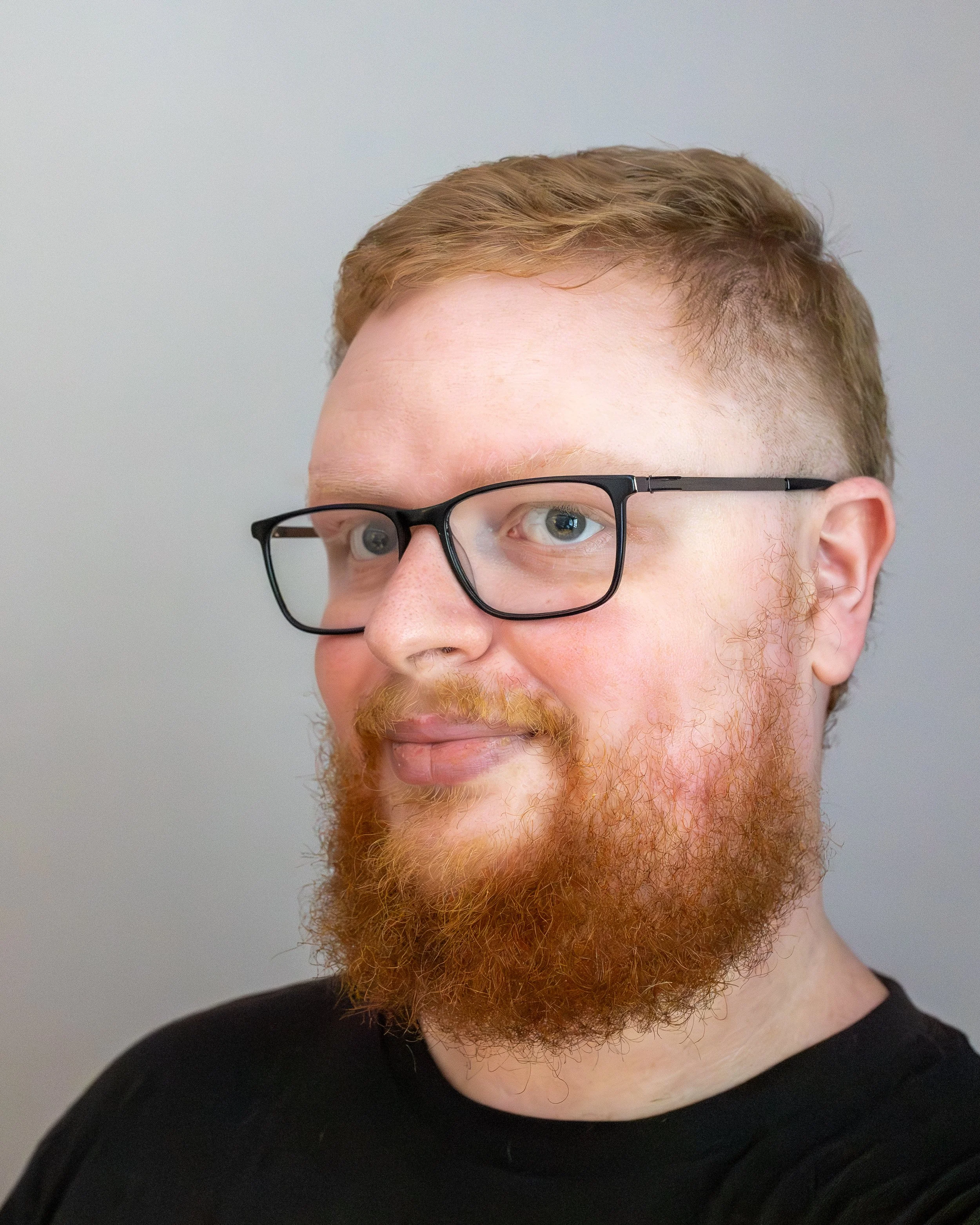 Close-up of a man with light skin, red hair, red beard, wearing black glasses, and dressed in a black shirt, against a plain gray background.