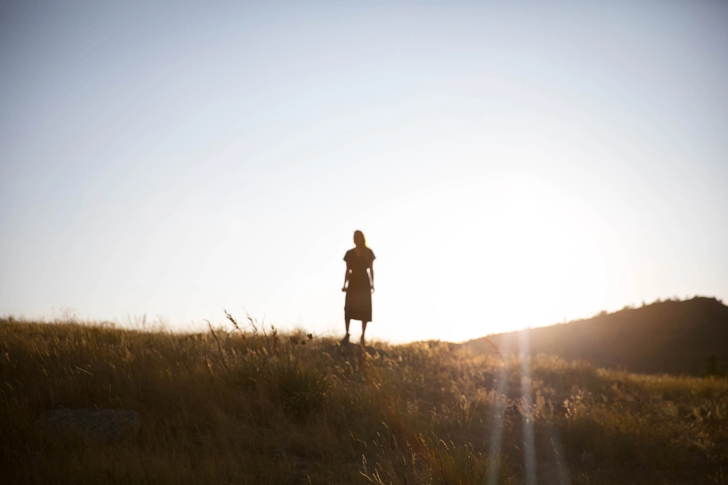 Silhouette of a woman standing on a grassy hill at sunset or sunrise with a clear sky and mountains in the background.
