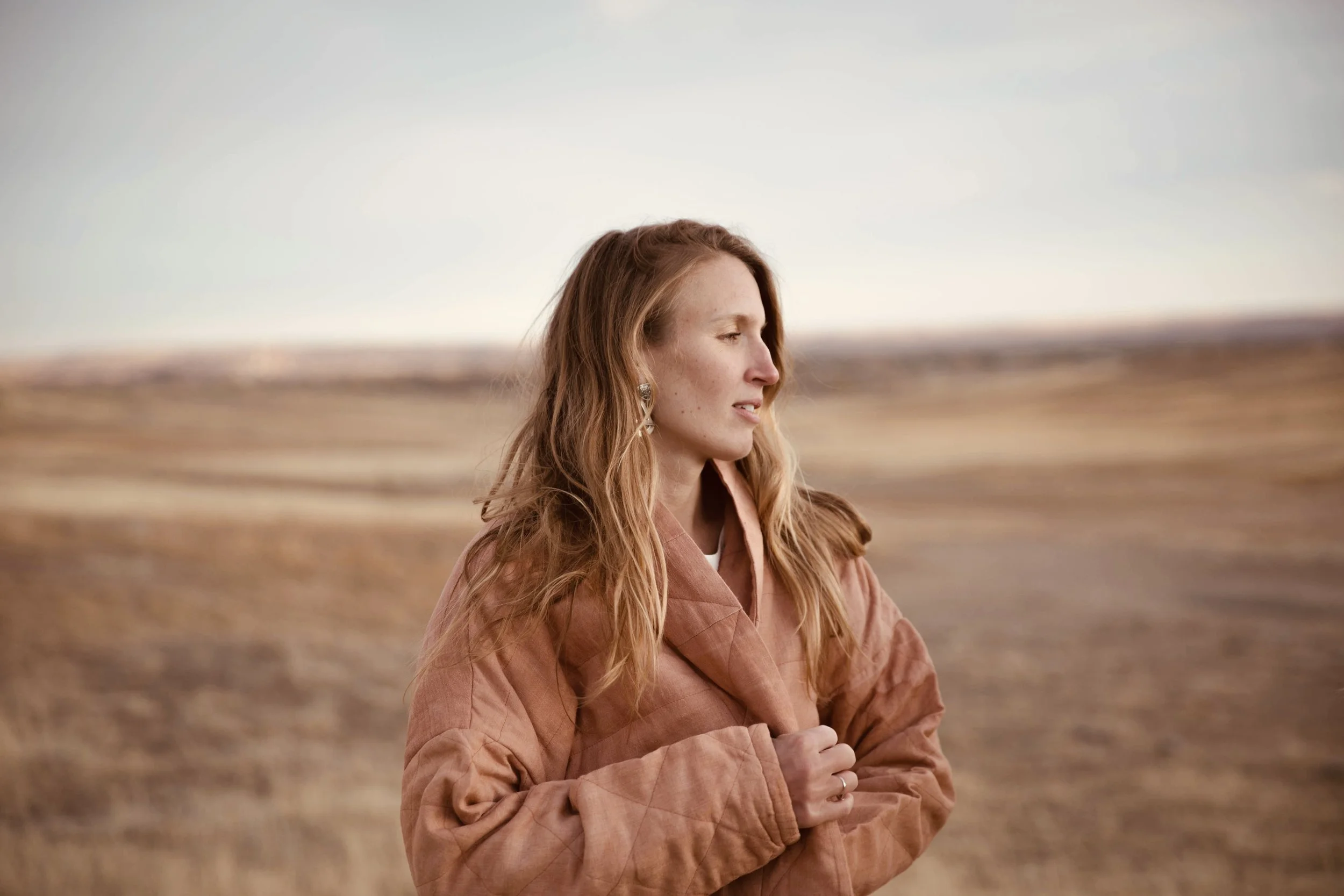 A woman with long red hair standing outdoors with her eyes closed, wearing a brown jacket, in a vast open field during the day.