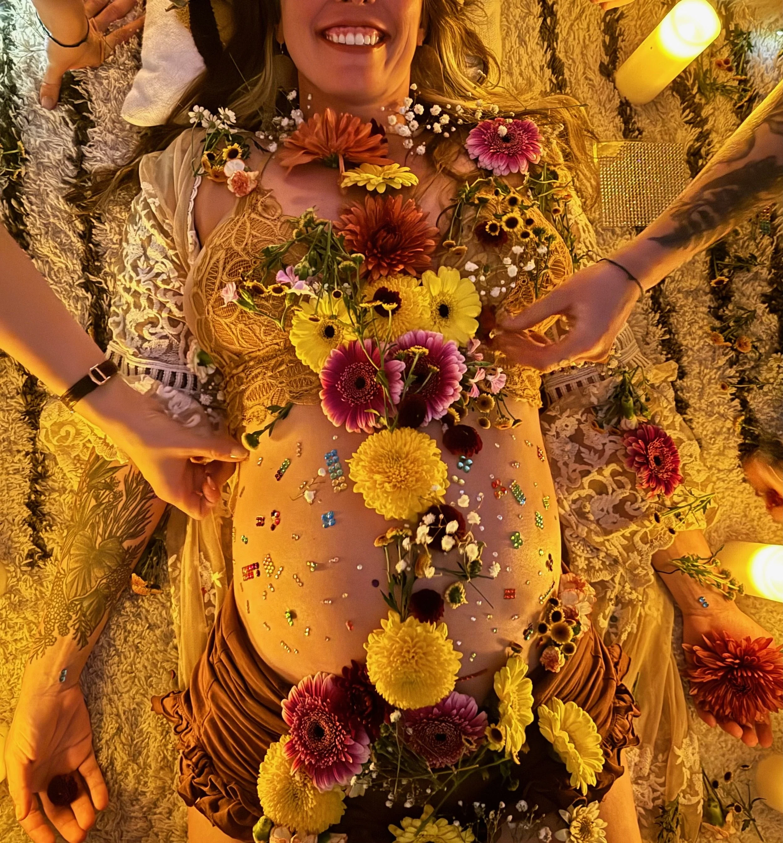 A woman lying on her back on a floral blanket, surrounded by flowers and candles, with a big smile on her face.