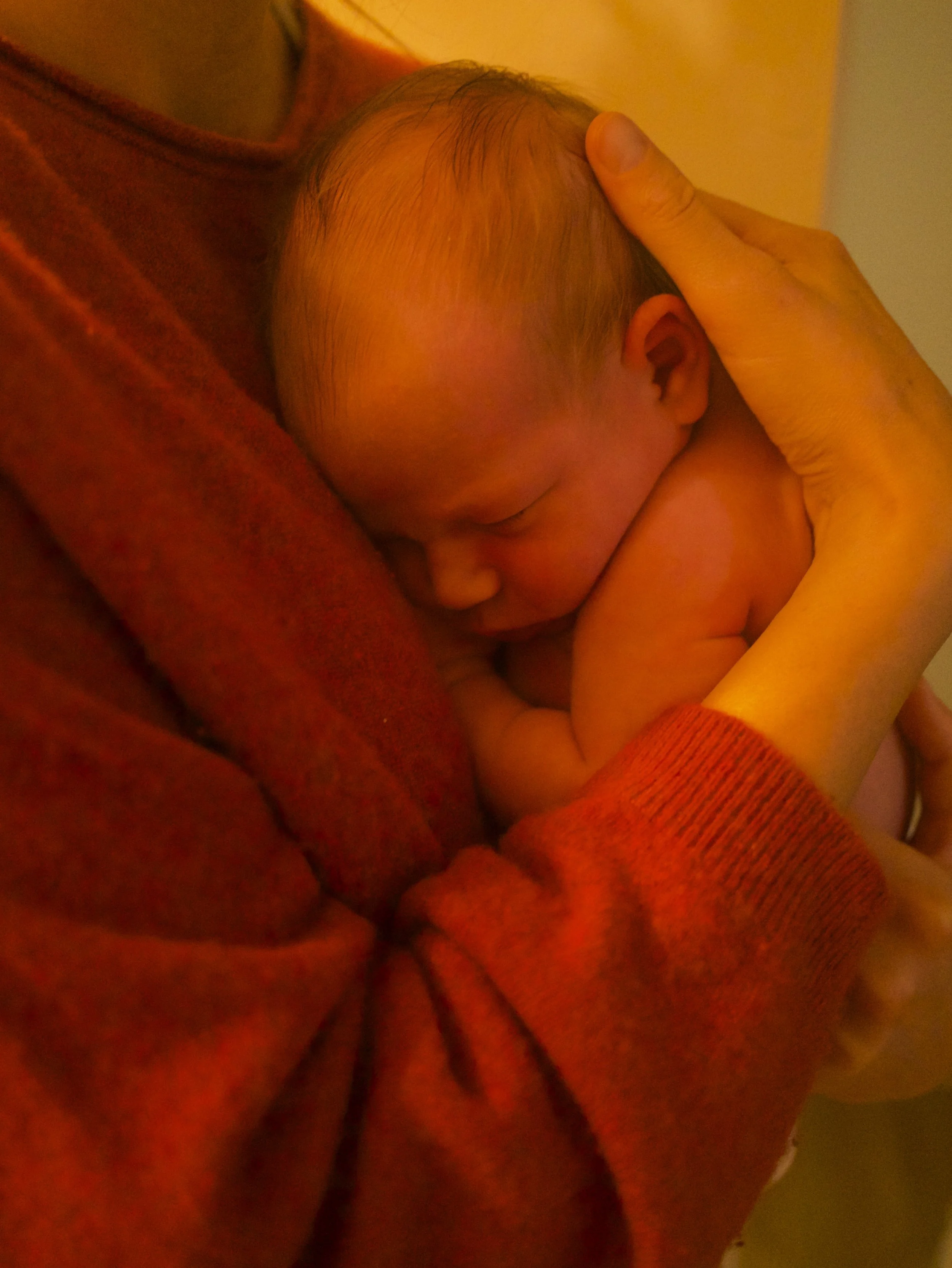 Close-up of a sleeping baby curled up, resting on an adult's arm and shoulder.