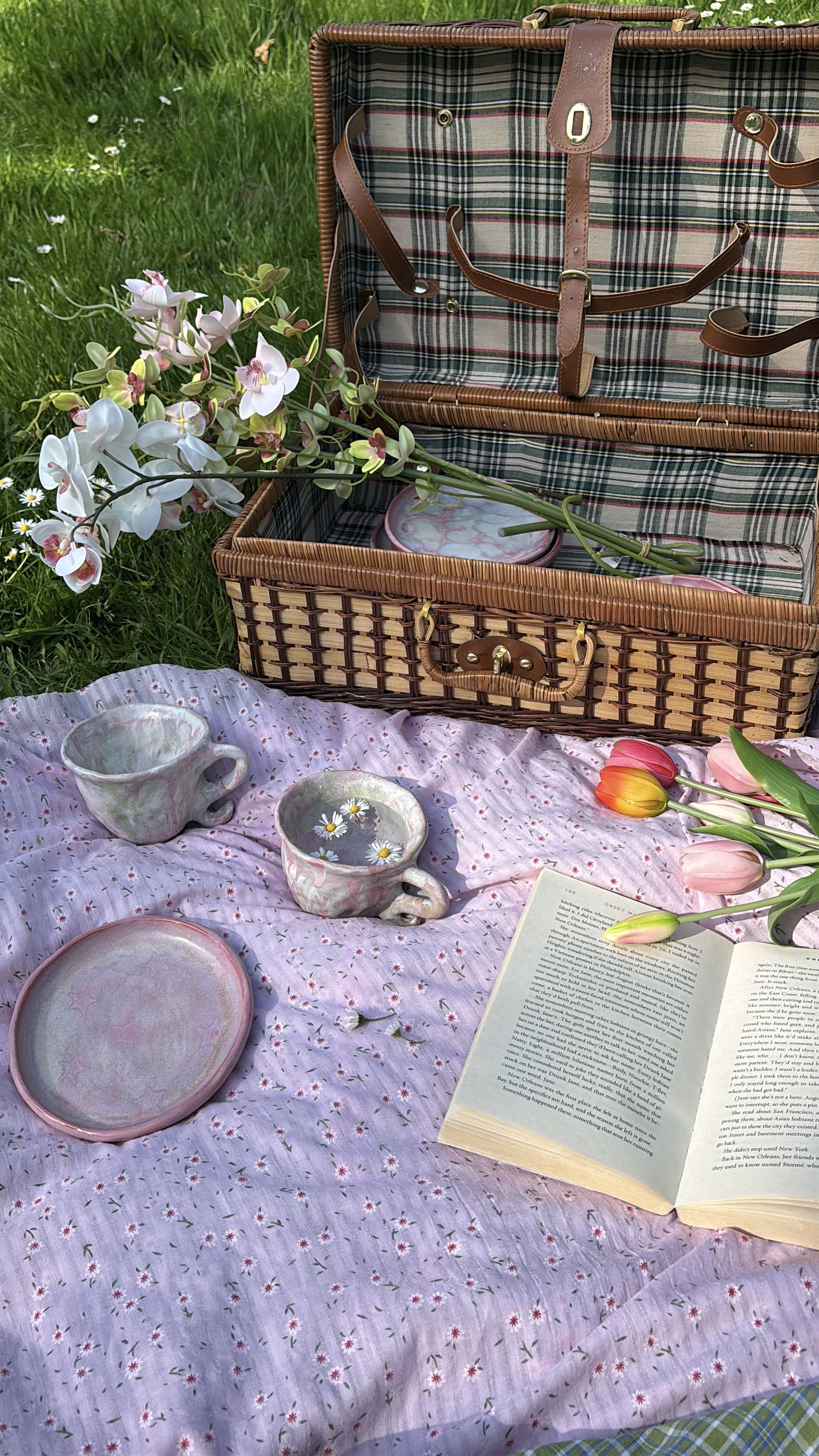 A picnic setup with a pink floral tablecloth, featuring two ceramic cups with daisies floating in water, a pink ceramic plate, and an open book. A wicker picnic basket with a plaid interior is open, holding a bouquet of white orchids. Pink tulips are also on the table, set outdoors on green grass with small white flowers.